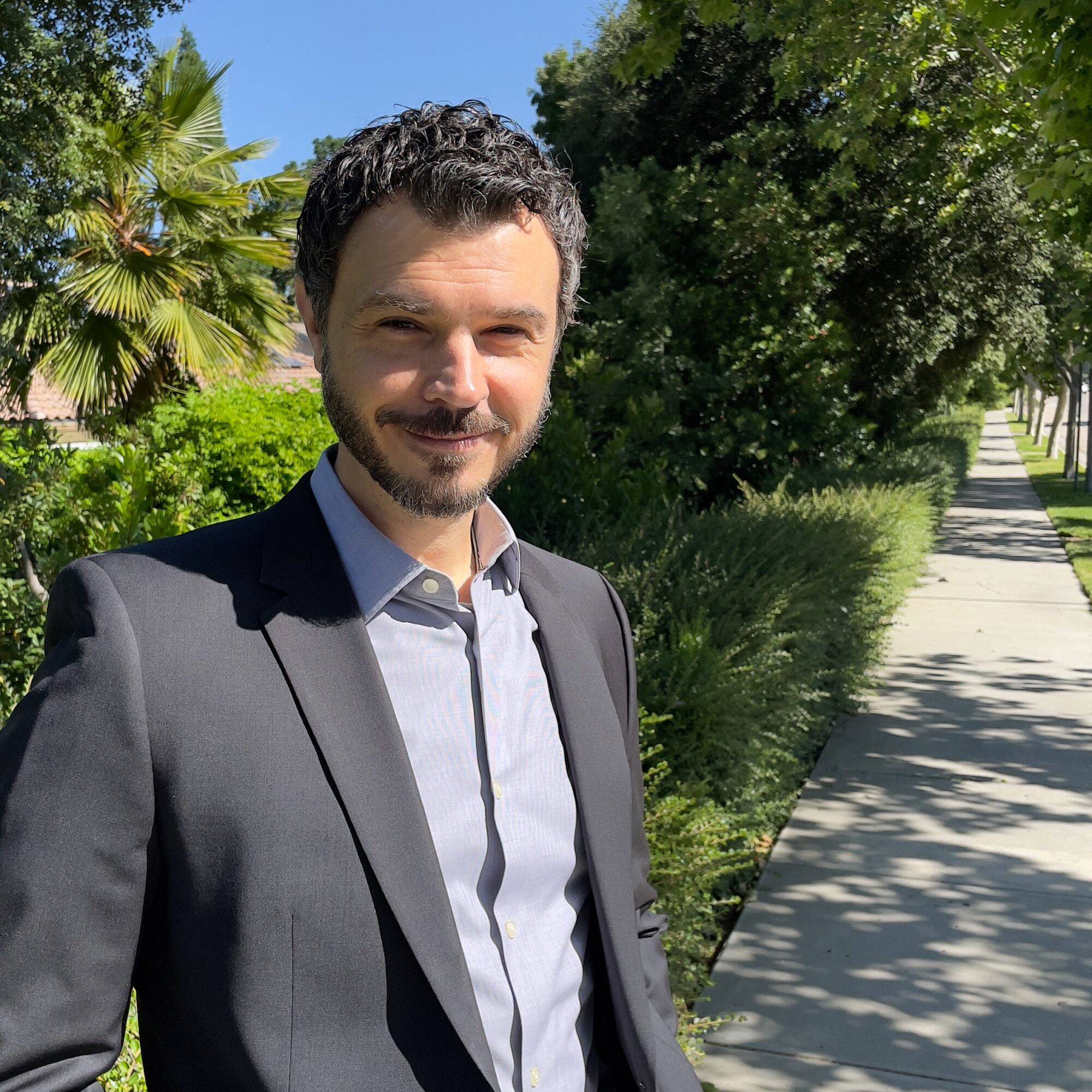 Man in suit standing outdoors on a sunny sidewalk with trees and greenery in the background.