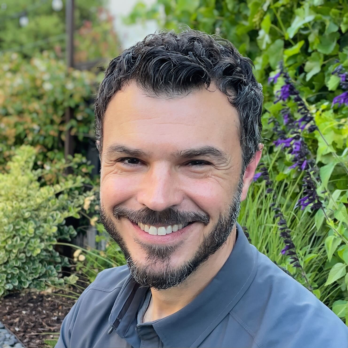 Man with dark curly hair and beard smiling outdoors with green plants and purple flowers in background