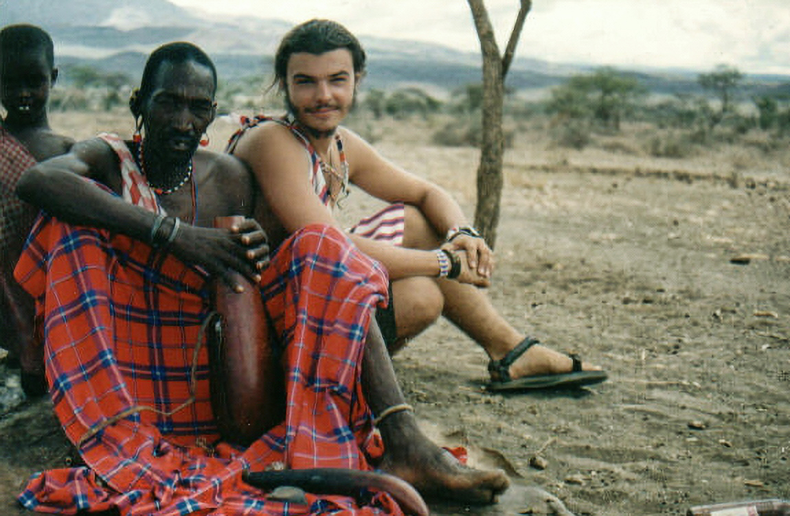 Two men sitting outdoors on the ground, one wearing a red plaid cloth, the other in a striped shirt, with a tree and landscape in background.