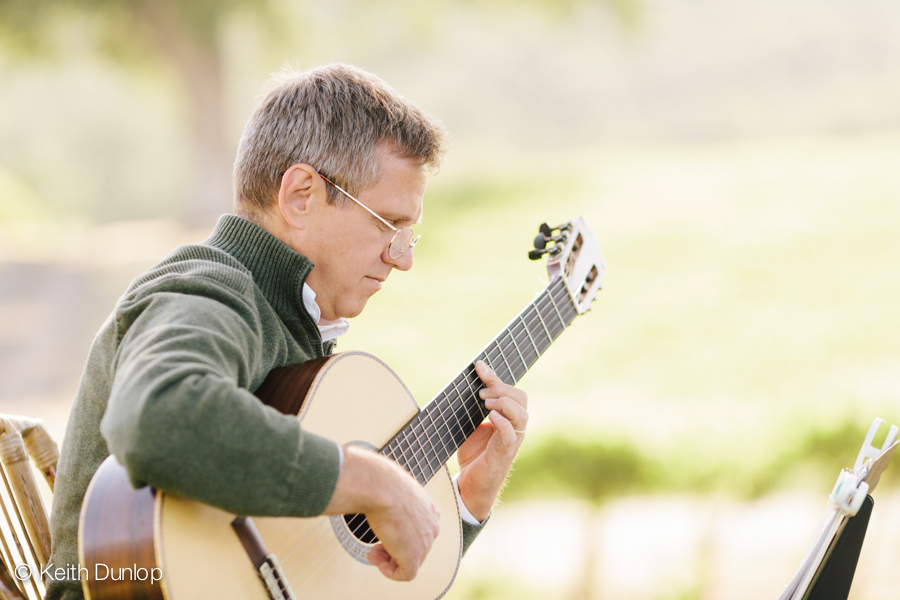 Man playing an acoustic guitar outdoors, wearing glasses and a green sweater, with a blurred natural background.