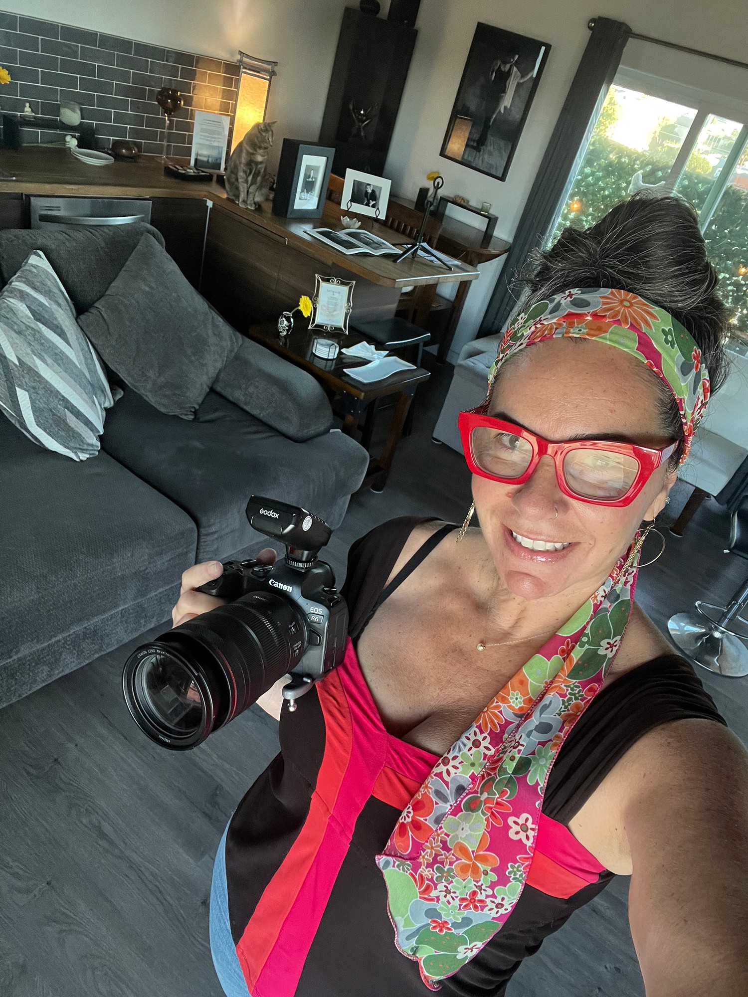 Woman with glasses and colorful headscarf smiling, holding a camera in a living room with a sofa and window.