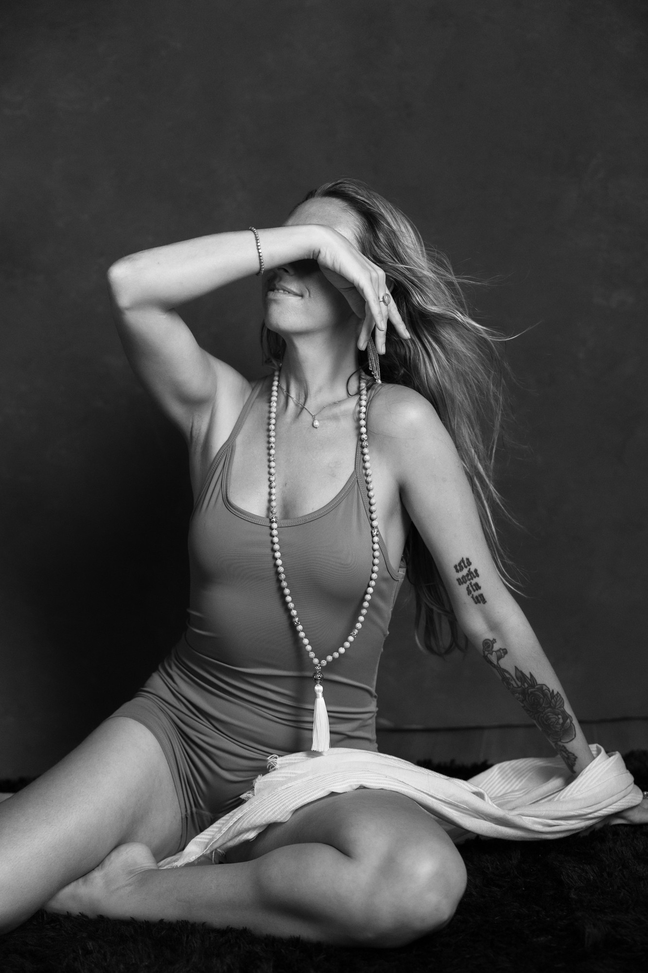 Woman sitting on the floor with long hair, wearing a tank top and a long necklace, covering her face with her hand.