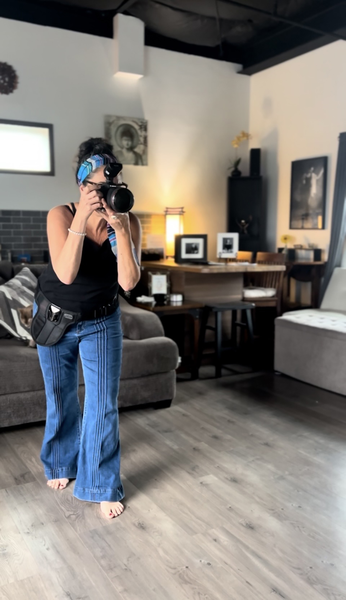 Person standing in a room holding a camera, wearing a black top and blue jeans, with a camera bag, in a cozy living space.