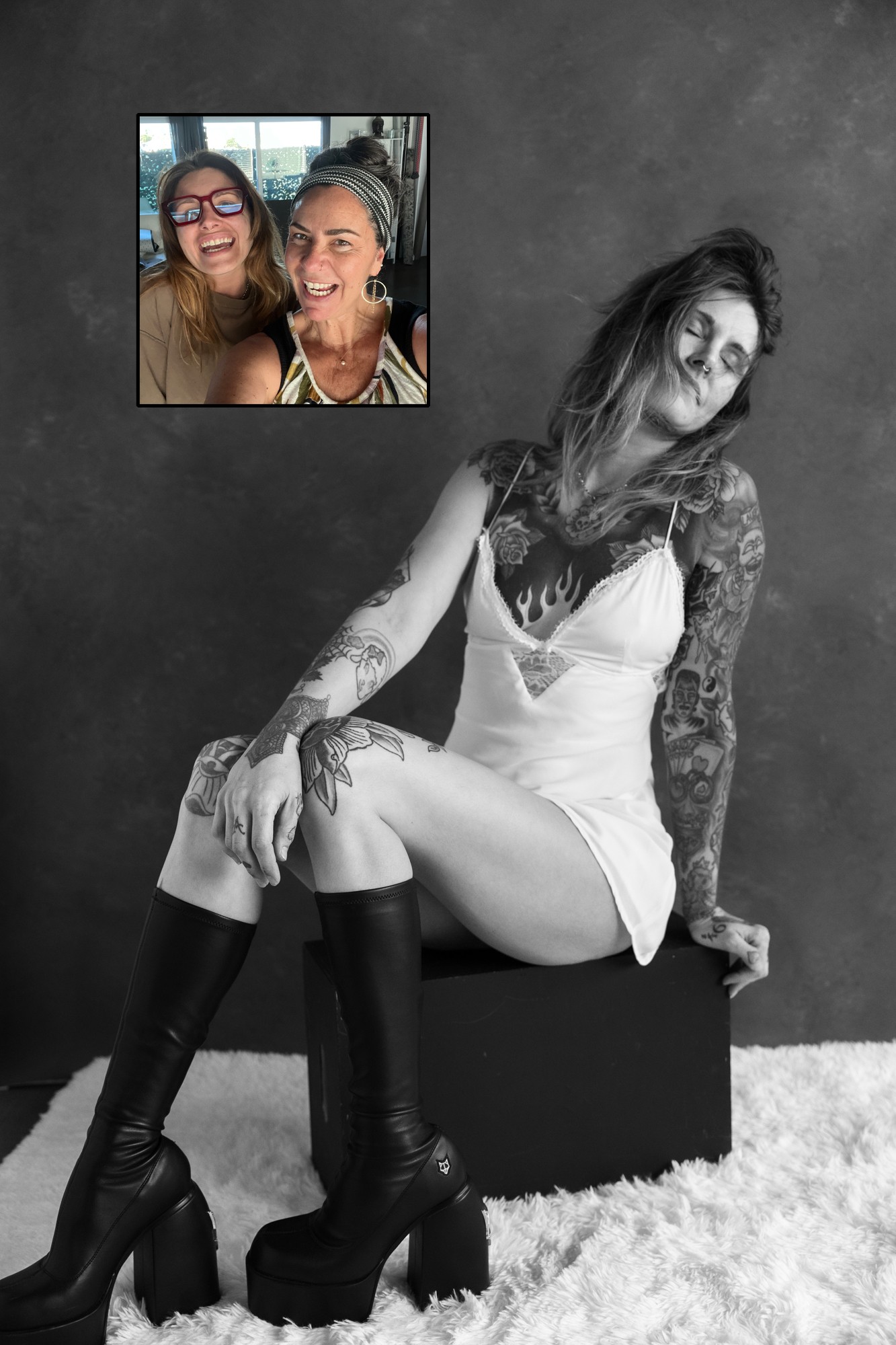 Black and white photo of a woman with tattoos sitting on a box, wearing a dress and high boots.