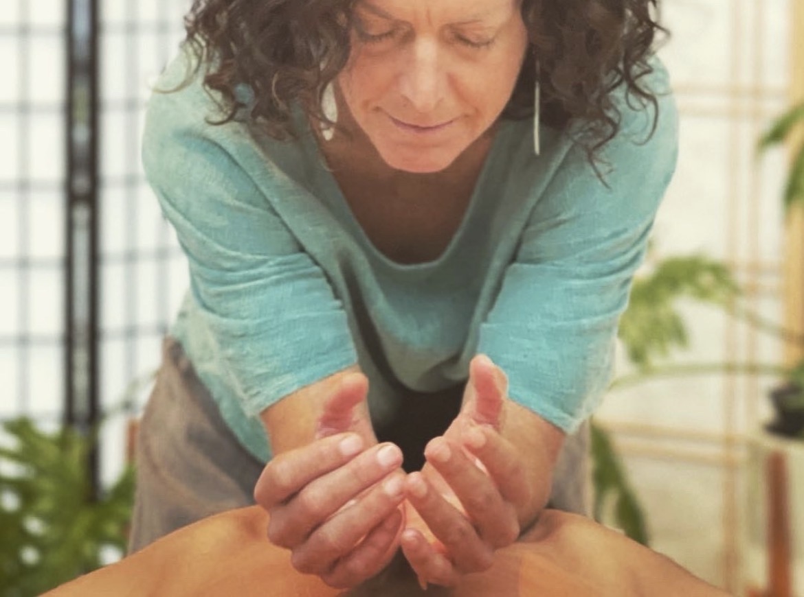 Woman with curly hair performs a massage or therapy on a person's hands in a bright room with plants.
