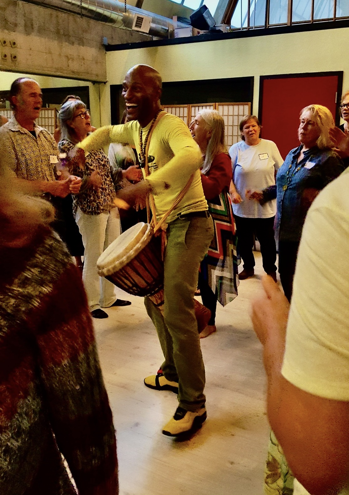 Person playing drum and dancing in a group indoors, with others watching and smiling.