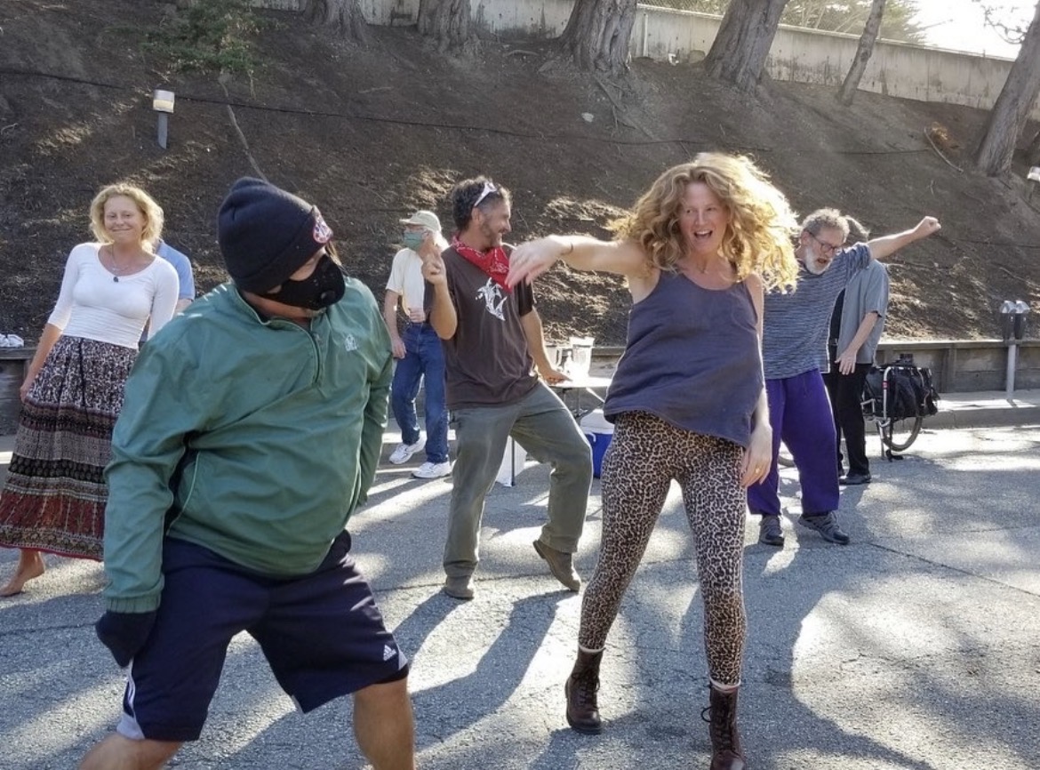 Group of people dancing outdoors on a sunny day, some with arms raised, in a park-like setting.