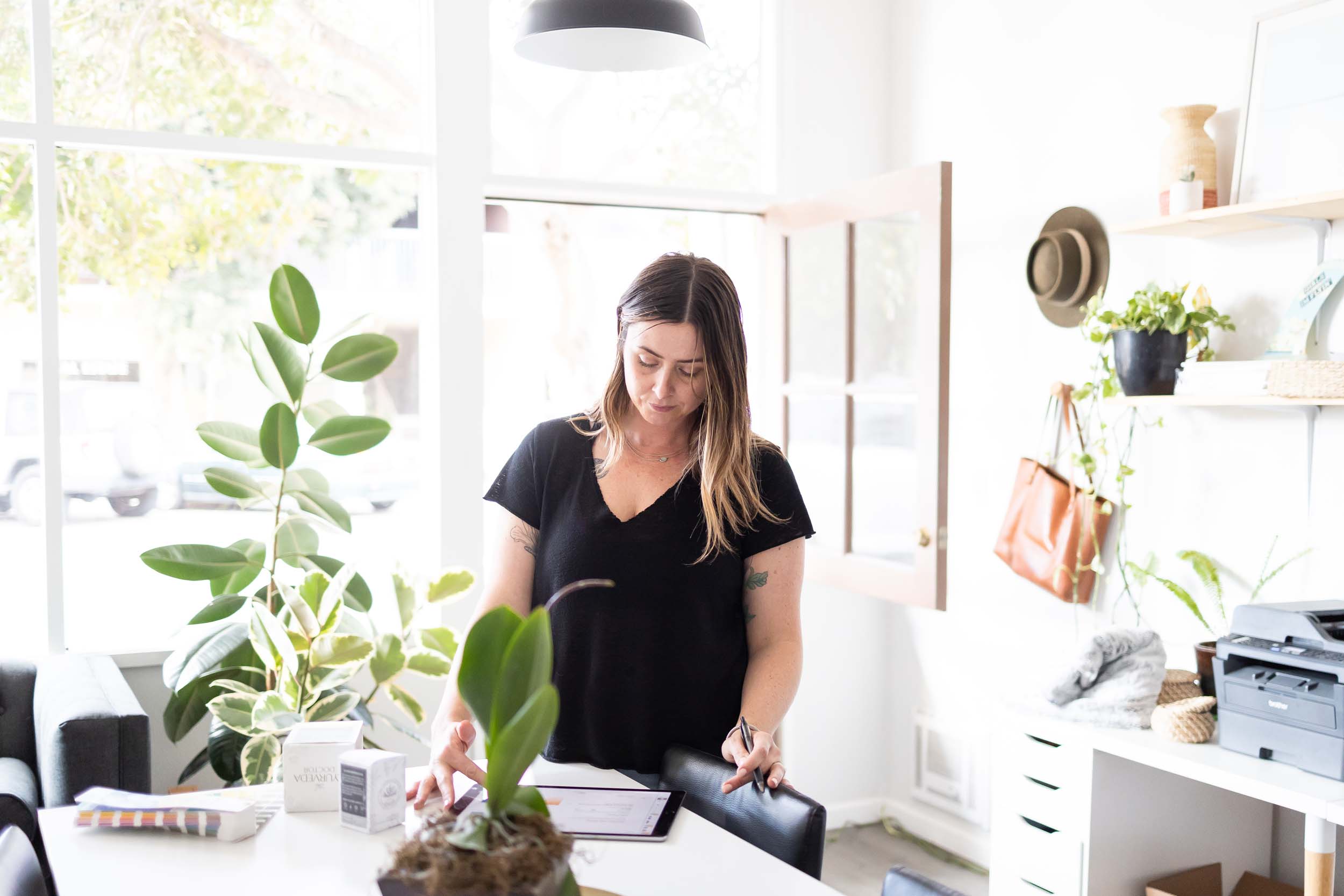 Woman with long hair in a black shirt standing near a window with plants and shelves, looking at a notebook.