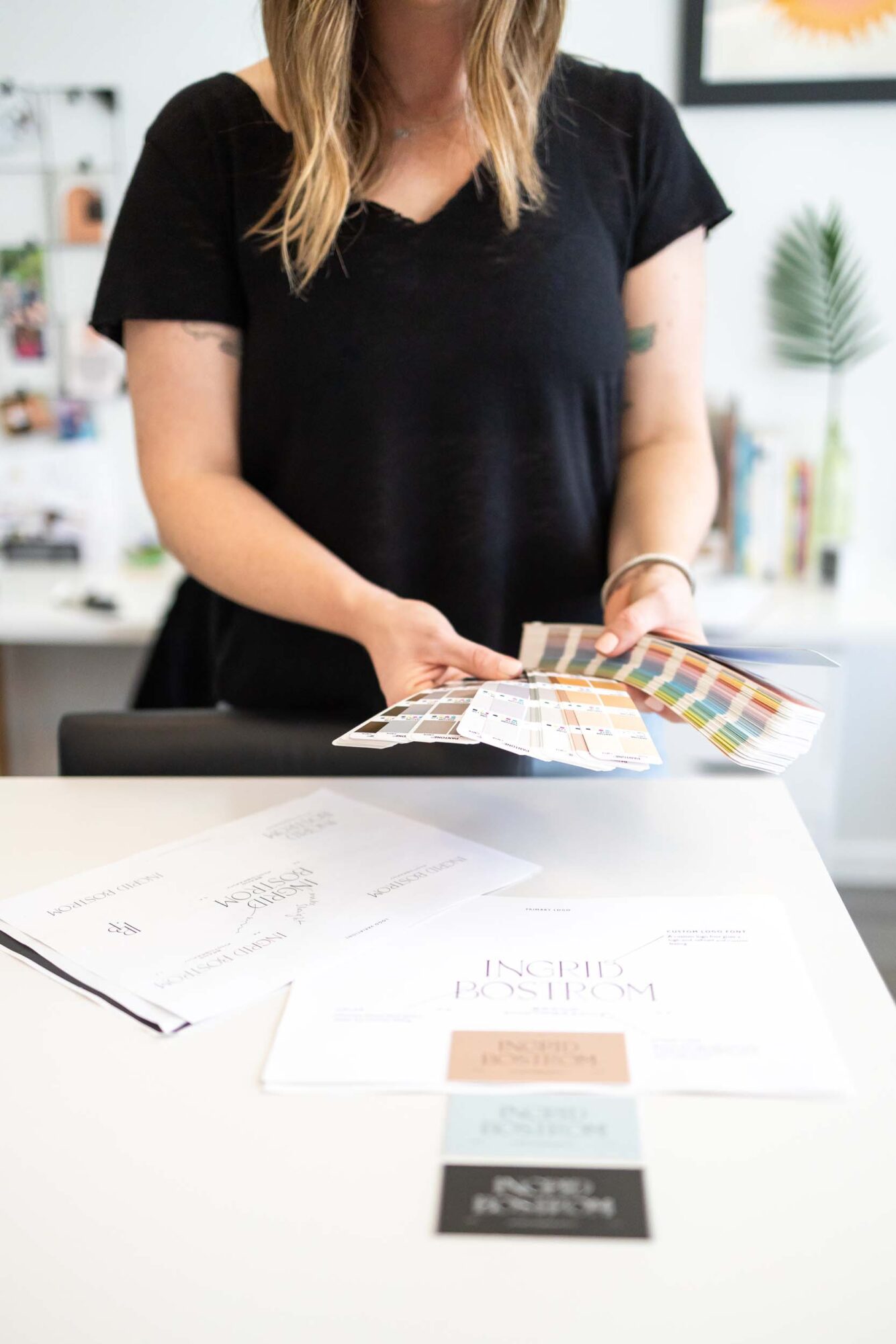 Person holding color swatches in a bright room with plants and artwork, standing at a table with papers.