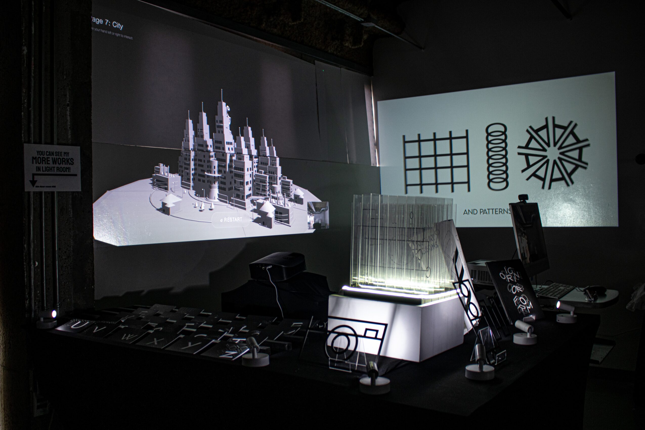 Exhibition with architectural models and digital displays, including cityscape models and geometric graphics on screens.