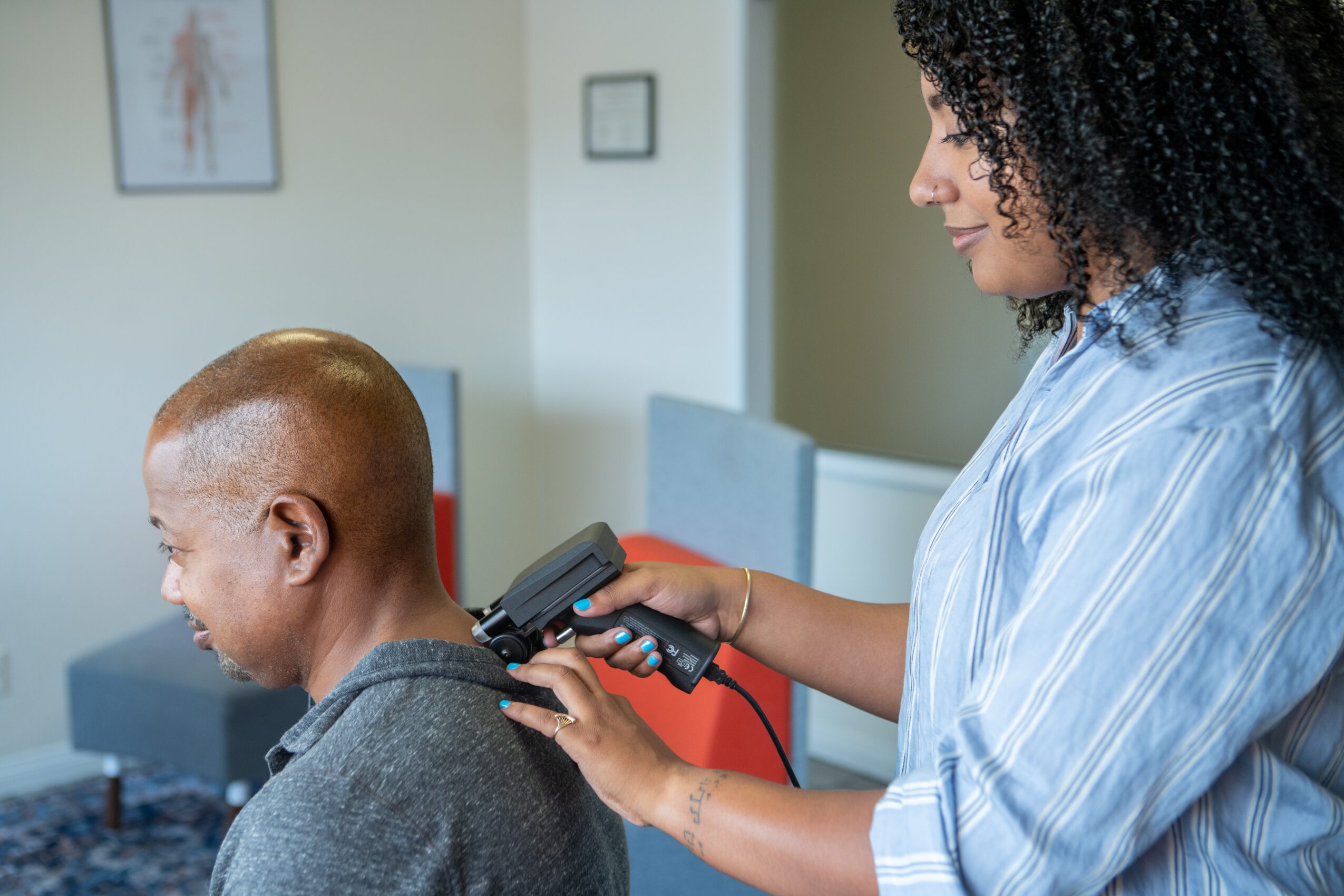 Woman using a device on a young person's head in a room with framed pictures, a computer, and chairs.
