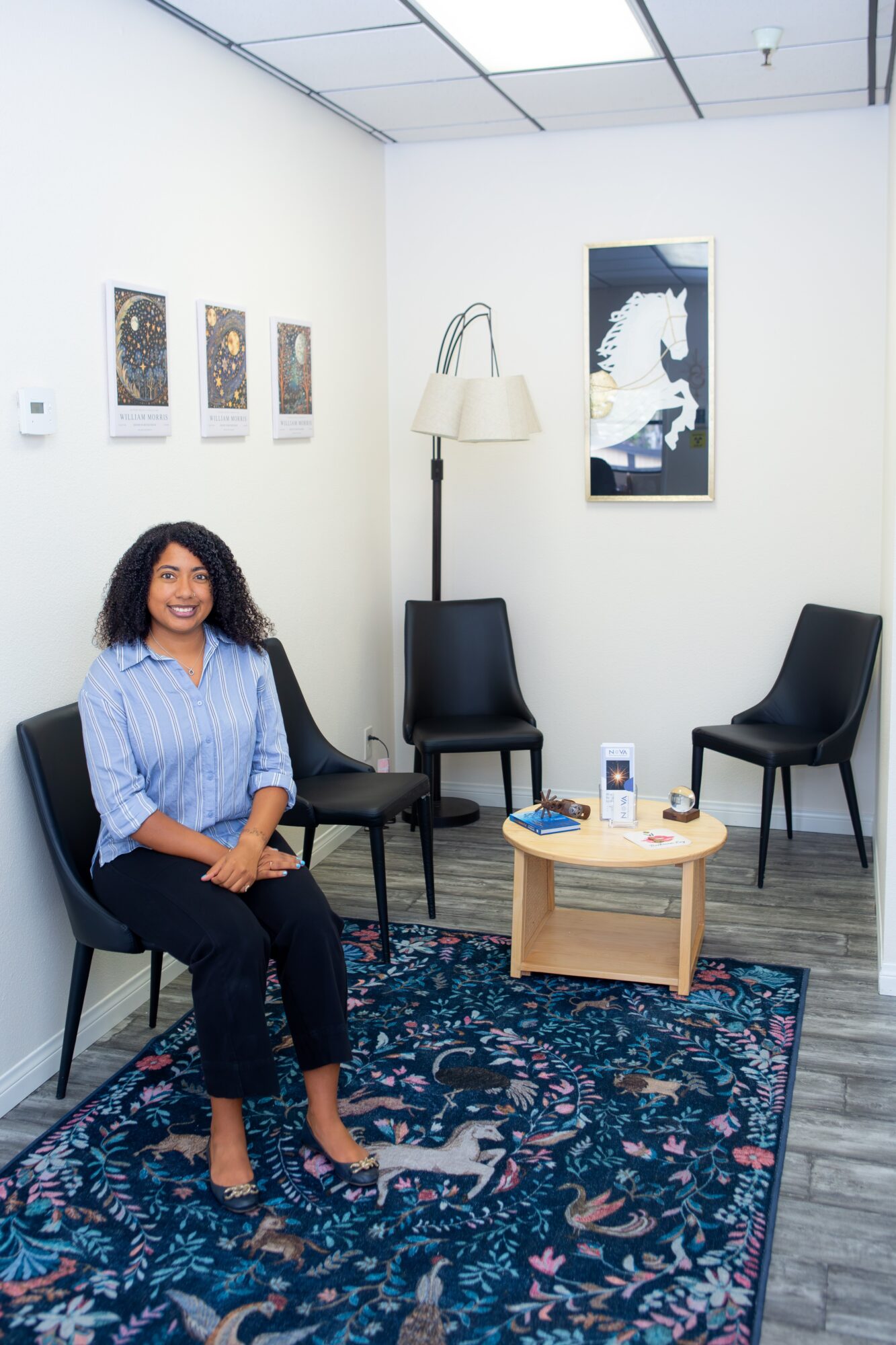 Smiling woman sitting on a black chair in a room with artwork, a lamp, and a small table, on a patterned rug.