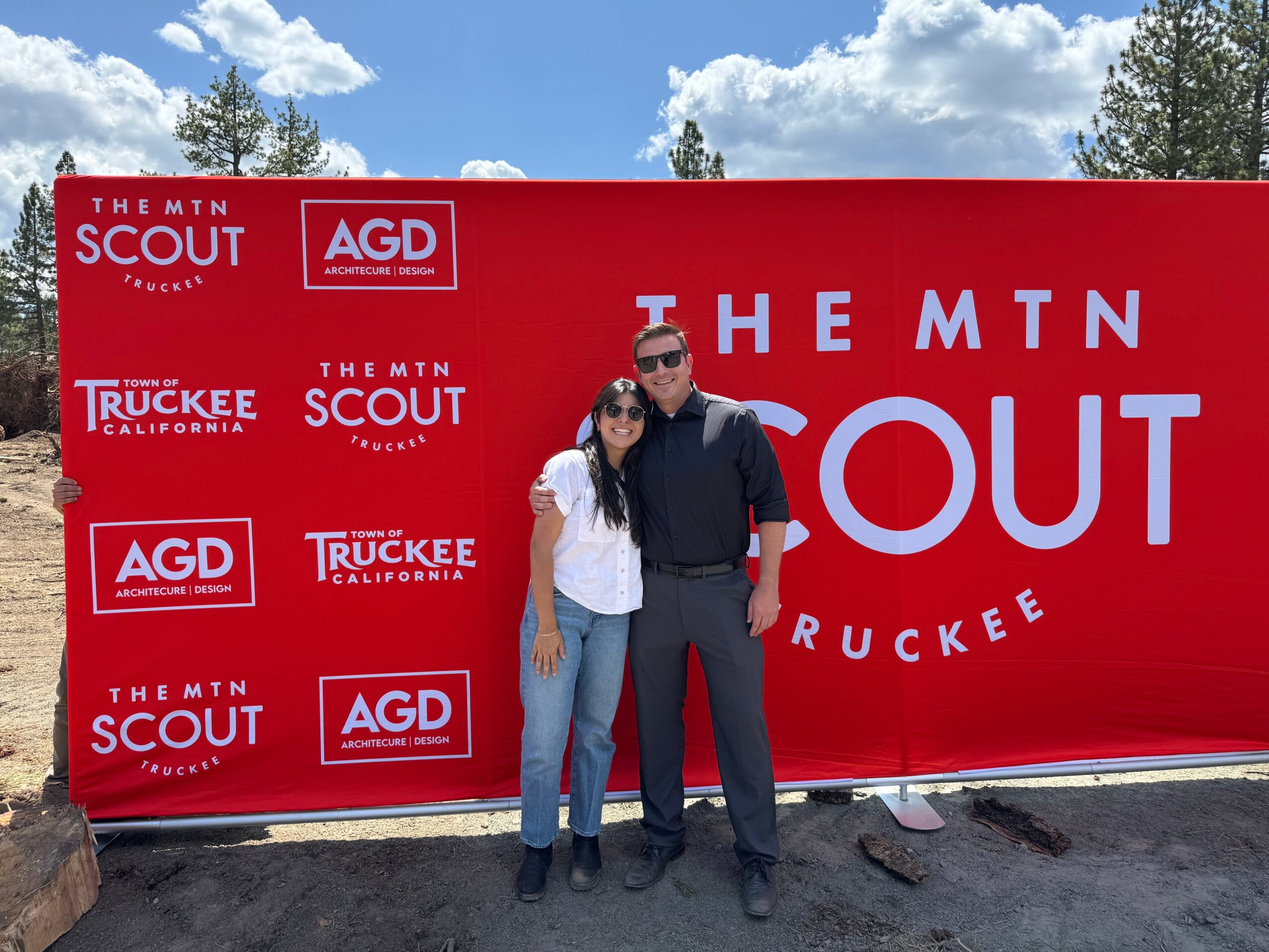 Two people standing in front of a large red sign with white text and logos, outdoors with trees and blue sky.