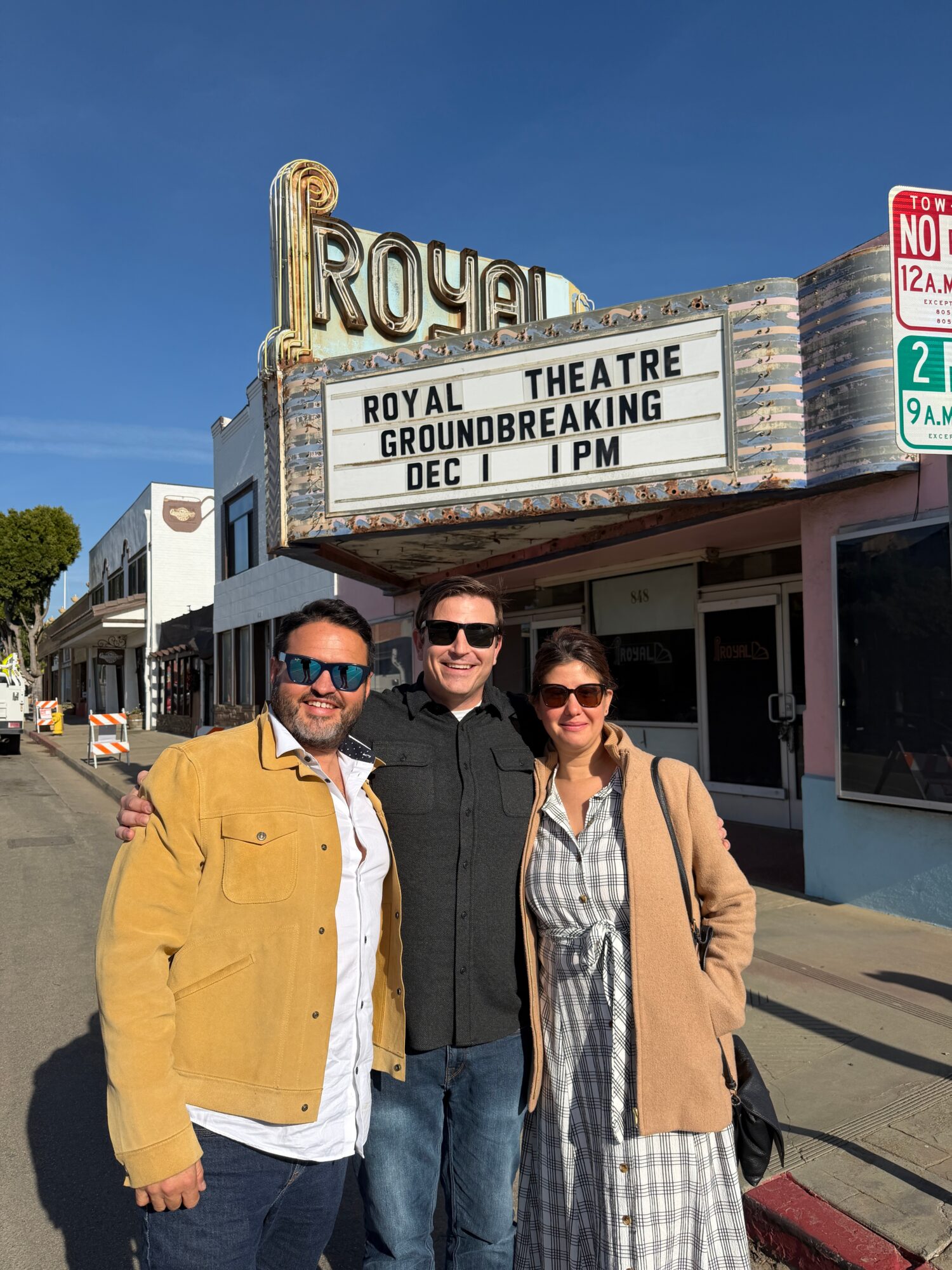 Three people stand in front of a theater with a marquee, smiling, with a clear blue sky above.