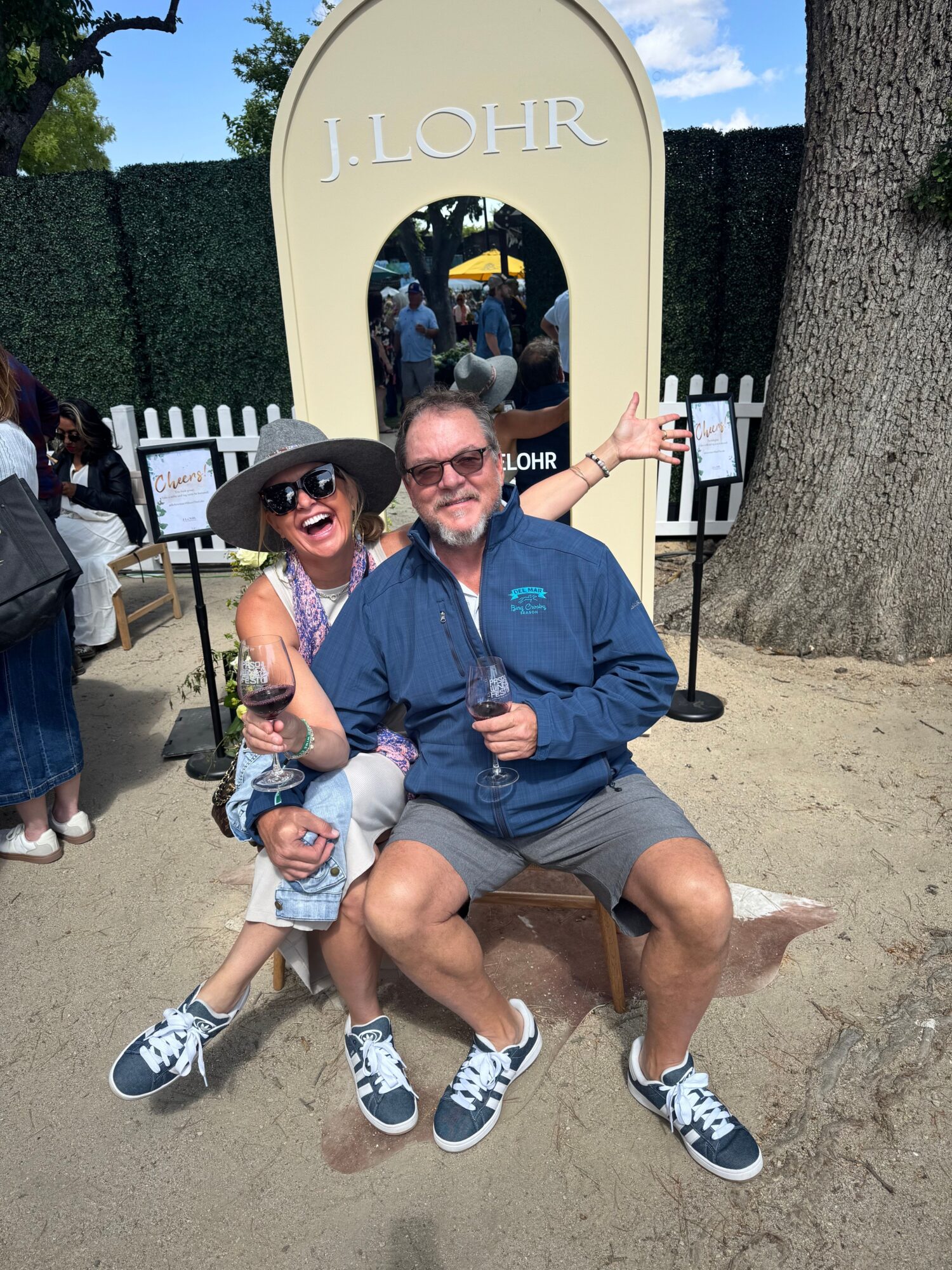 Two people sitting on a bench at a park, one with a stroller, smiling, with trees and a fence in the background.