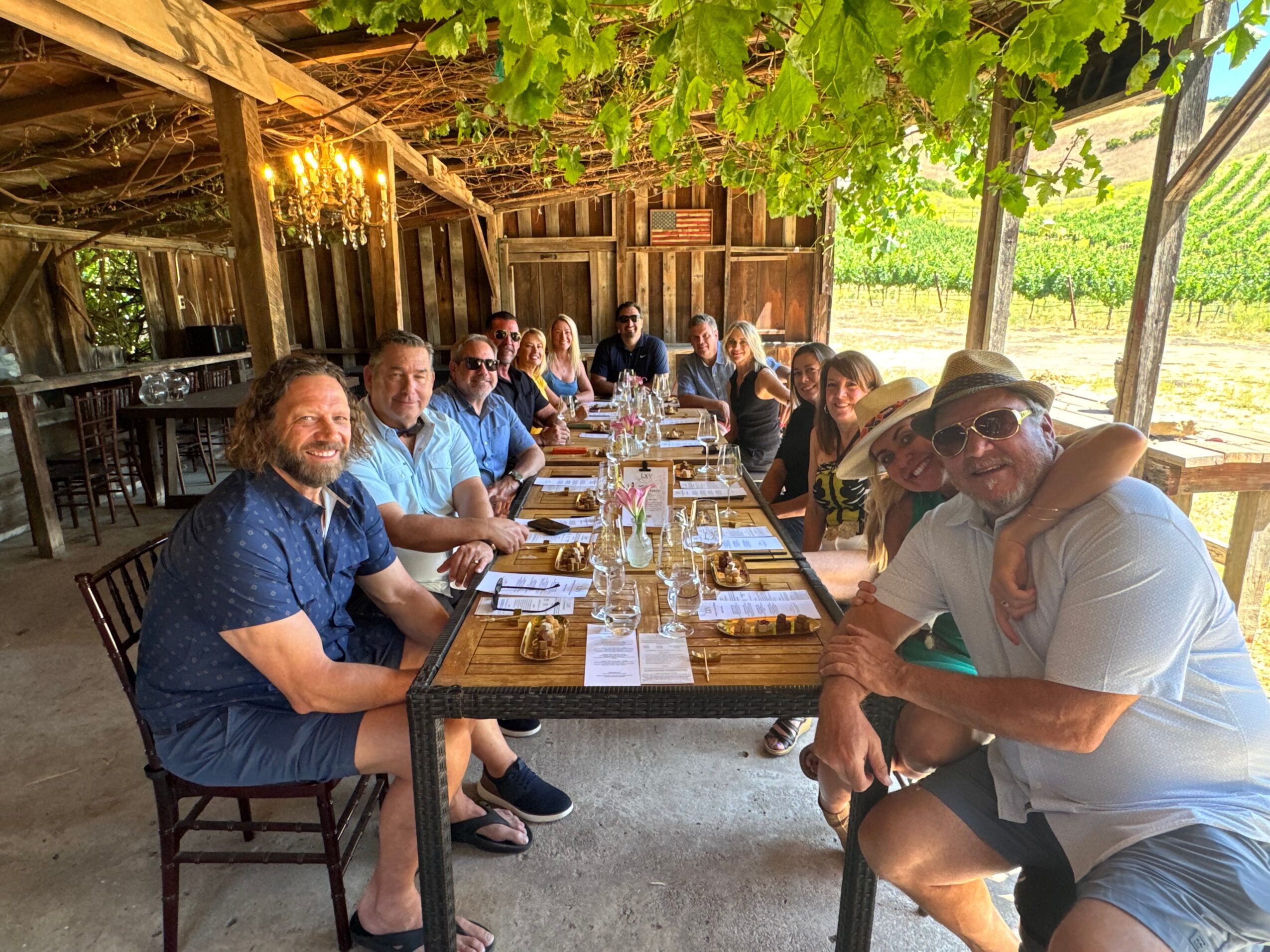 Group of people sitting at a long outdoor table under a tree, enjoying a meal together.