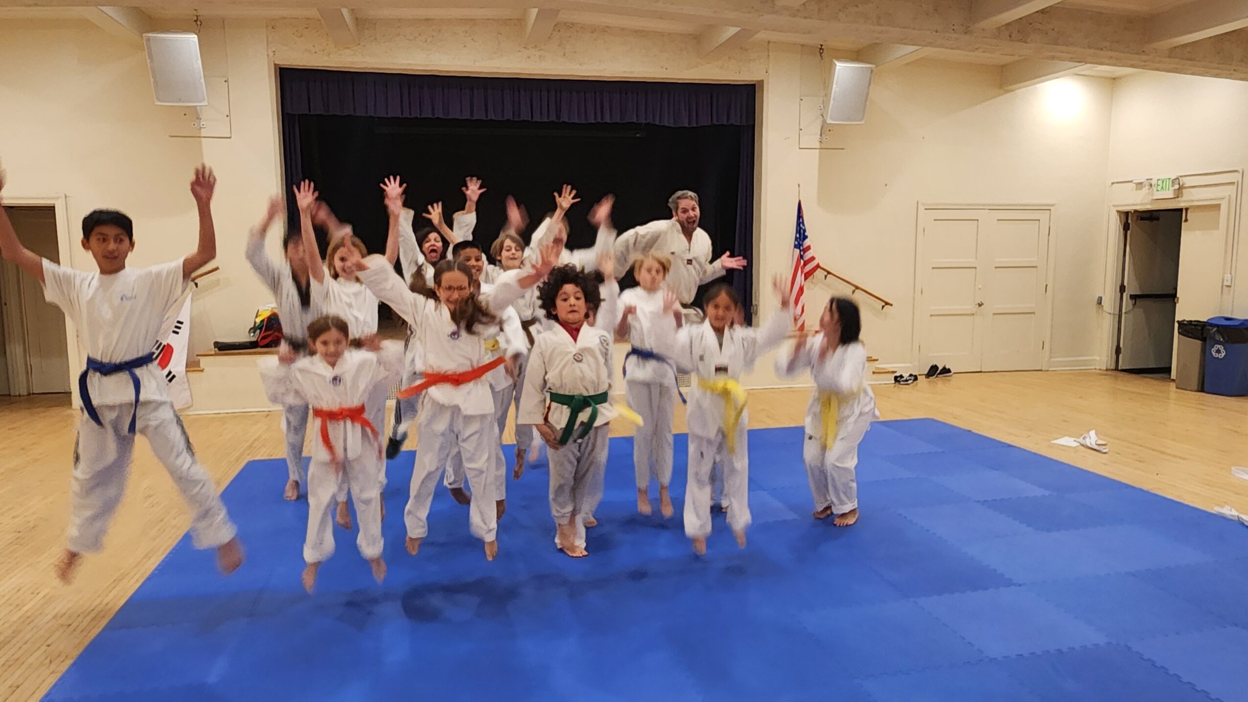 Group of children in martial arts uniforms with colored belts, standing on a blue mat in a gymnasium, raising their hands.