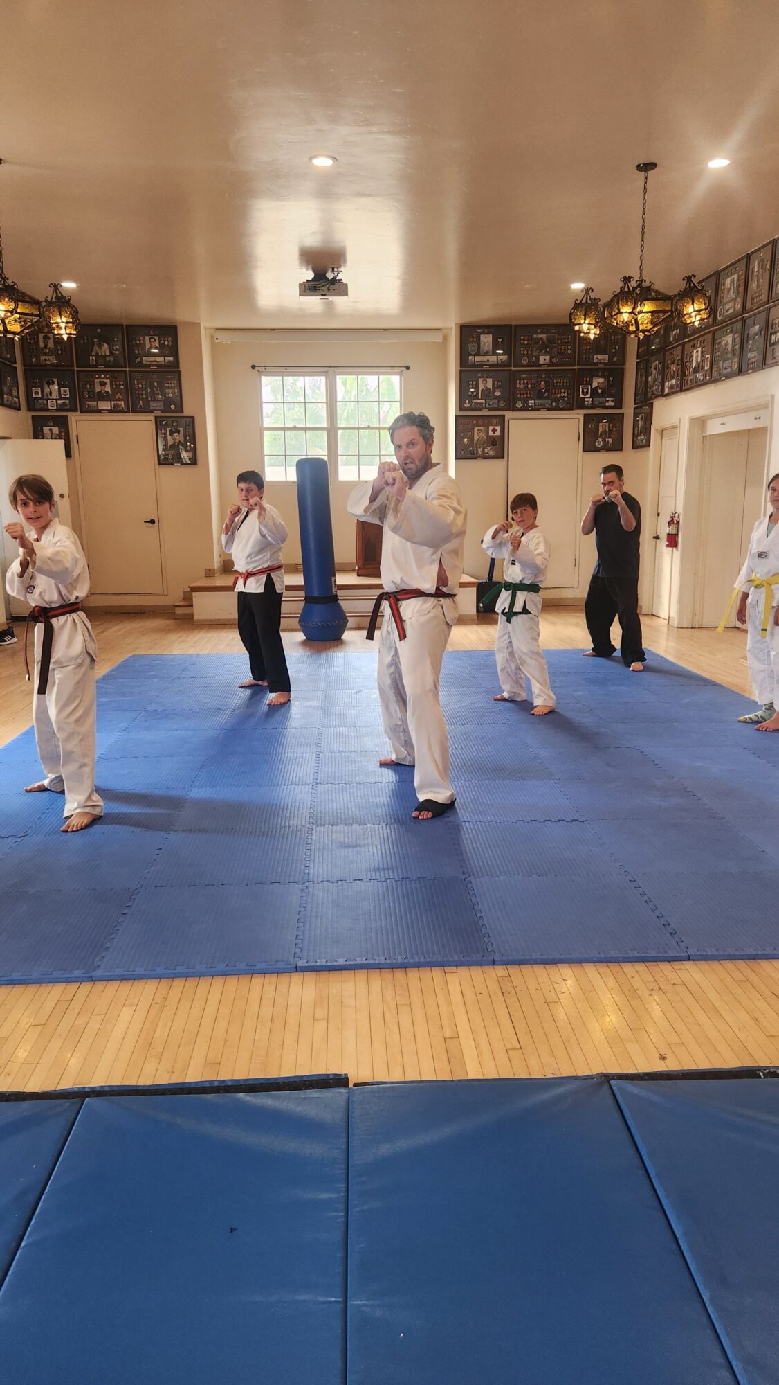 People practicing martial arts on a blue mat in a room with large windows and trophies, with a person in a white uniform in the foreground.