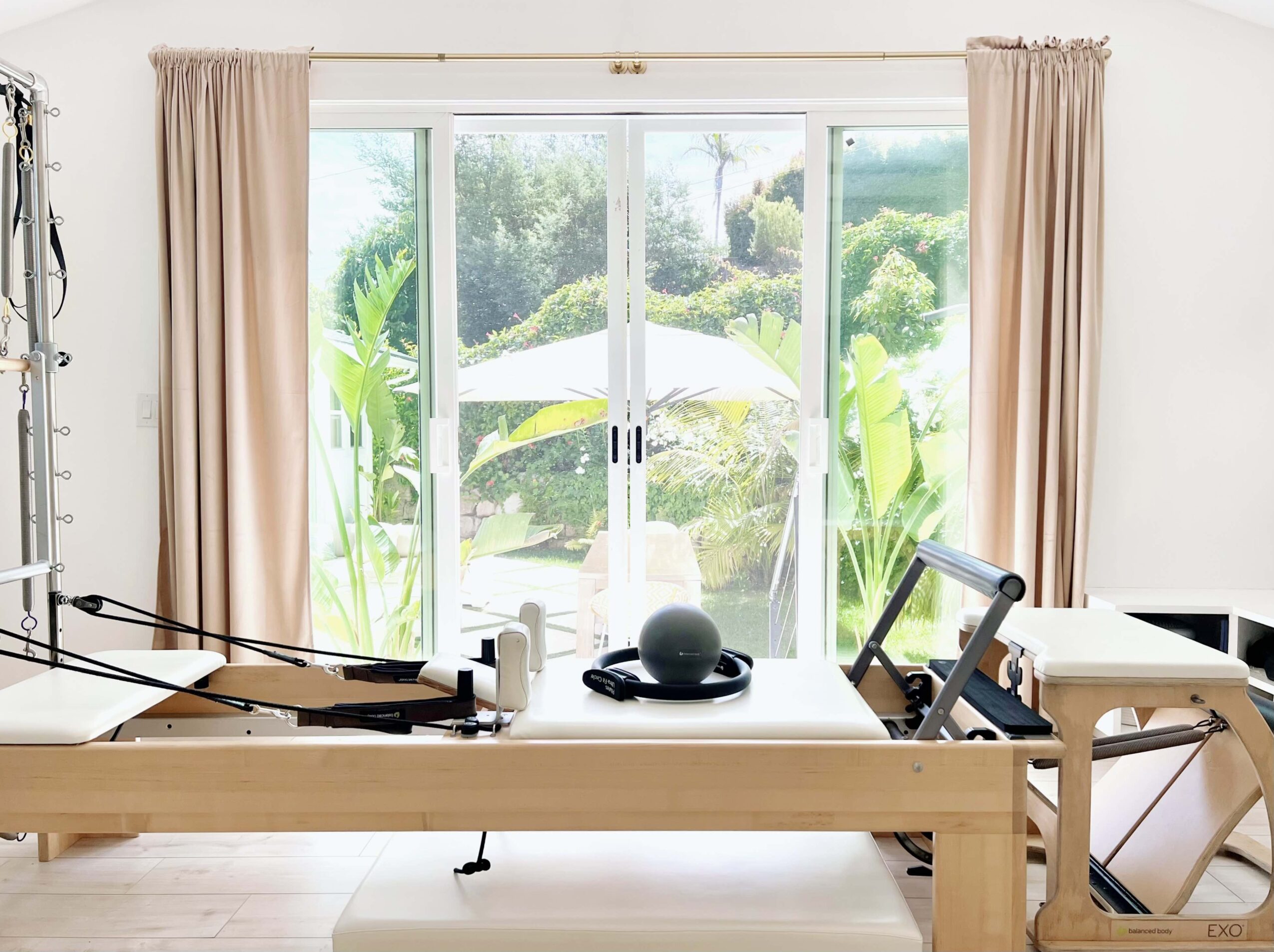Indoor room with large sliding glass doors opening to a garden with greenery and an umbrella, beige curtains, and exercise equipment on a wooden table.