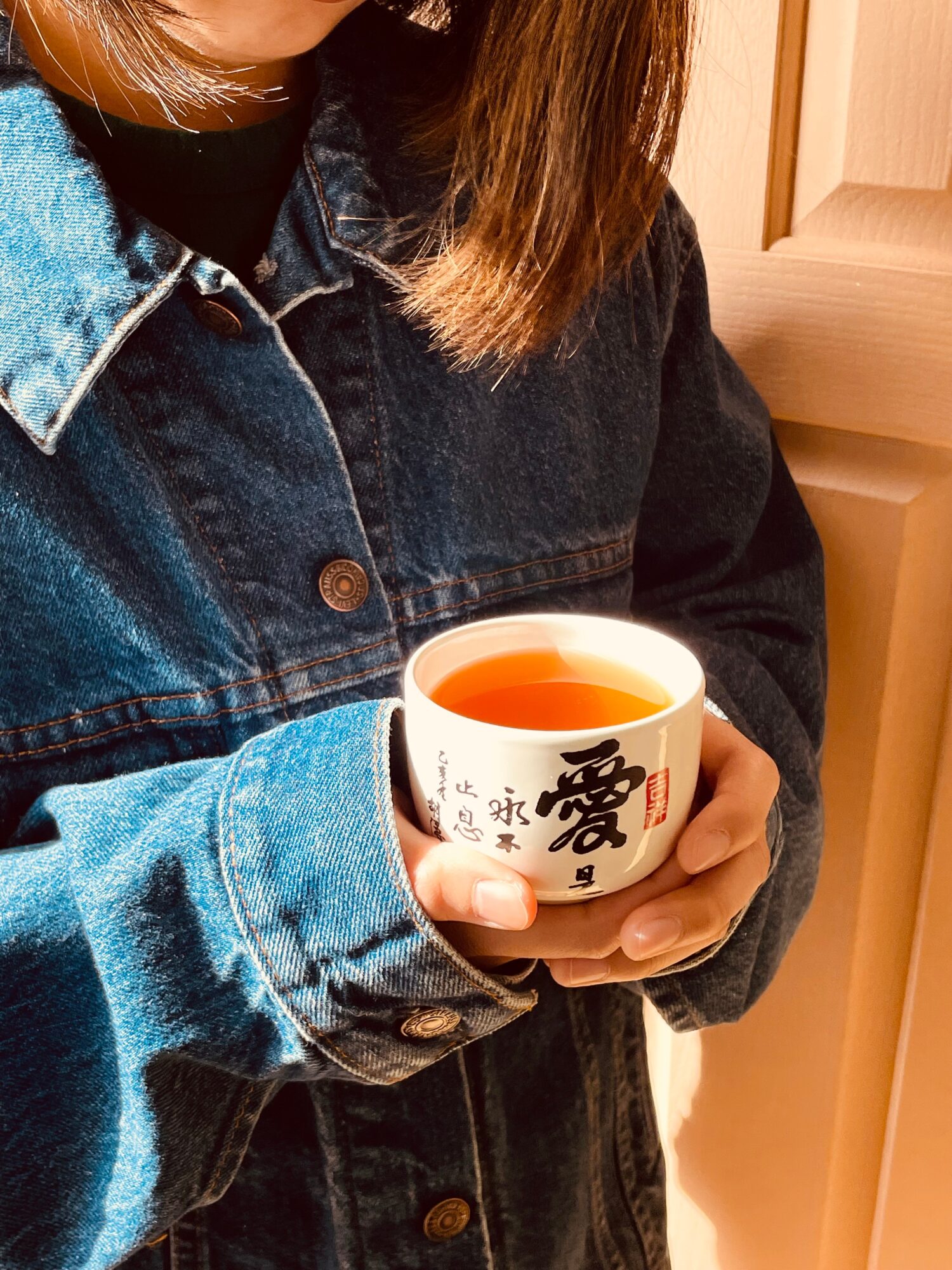 Person holding a white cup with black and red characters, wearing a denim jacket, indoors.