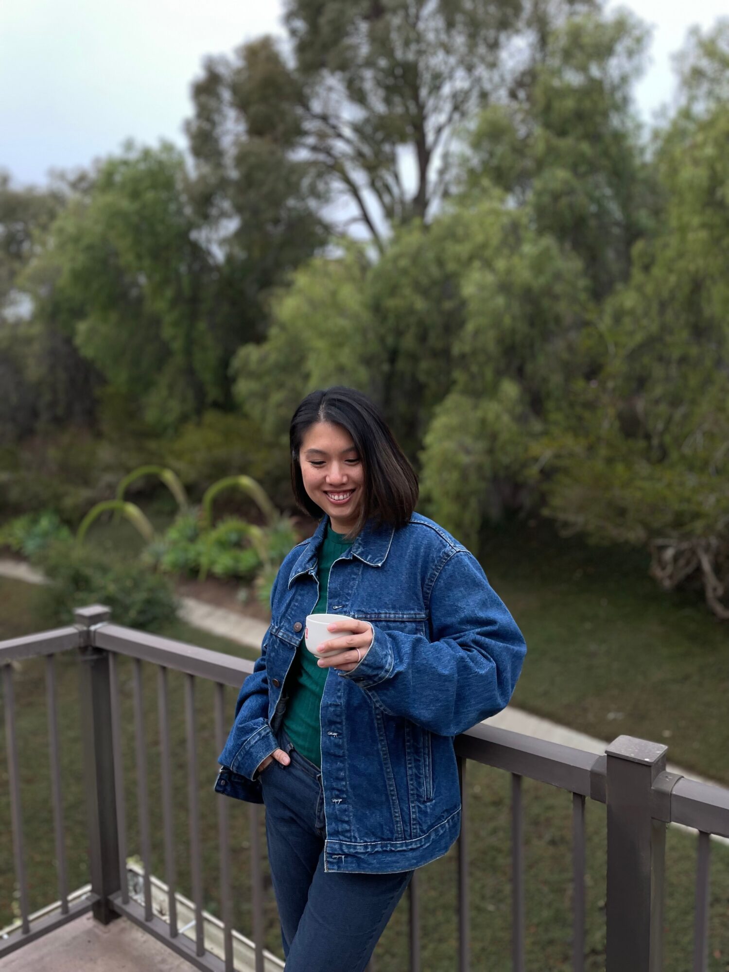 Young woman with shoulder-length dark hair smiling, holding a cup, standing on a wooden bridge with trees in the background.