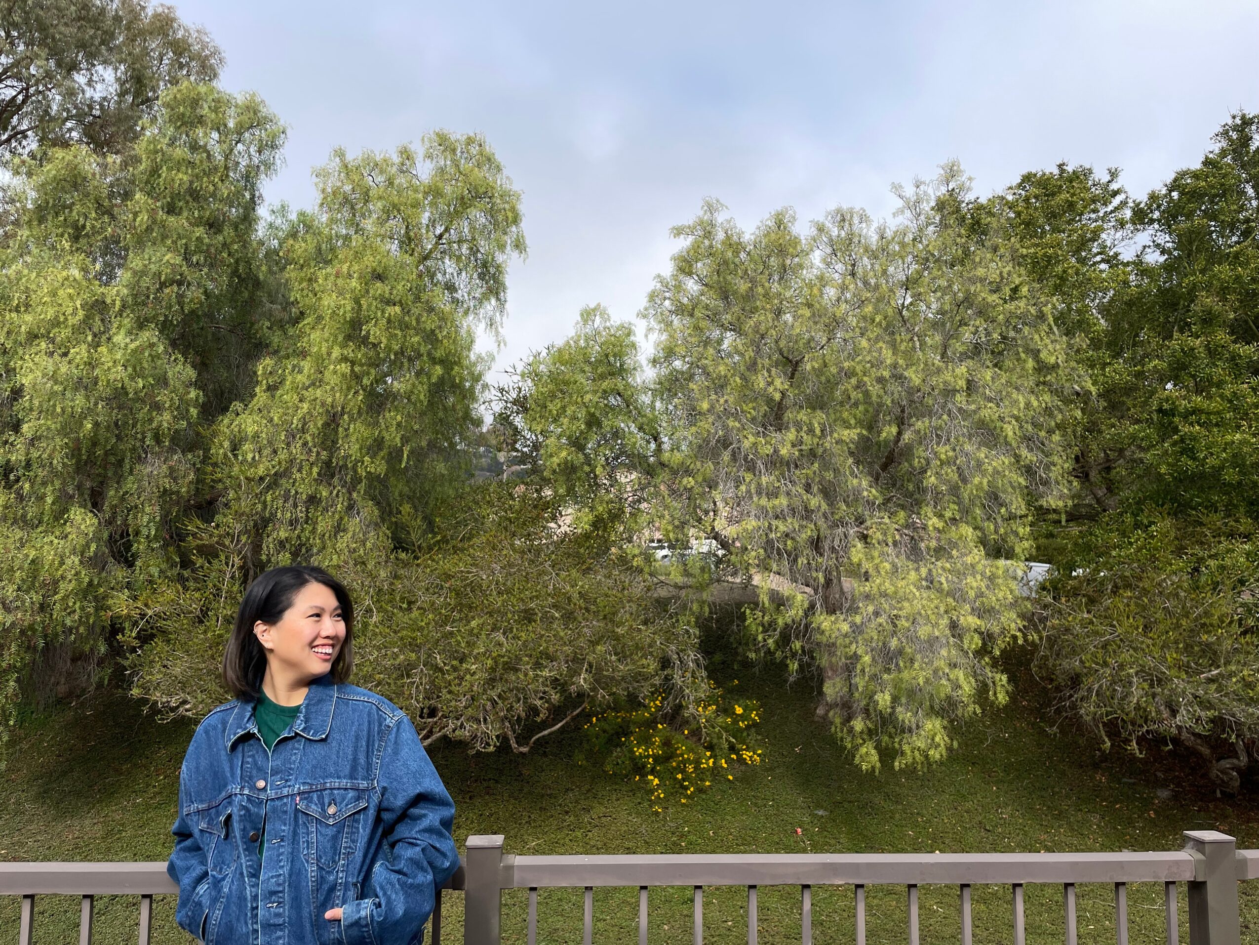 Woman in denim jacket standing outdoors near a wooden fence with trees and greenery in the background.
