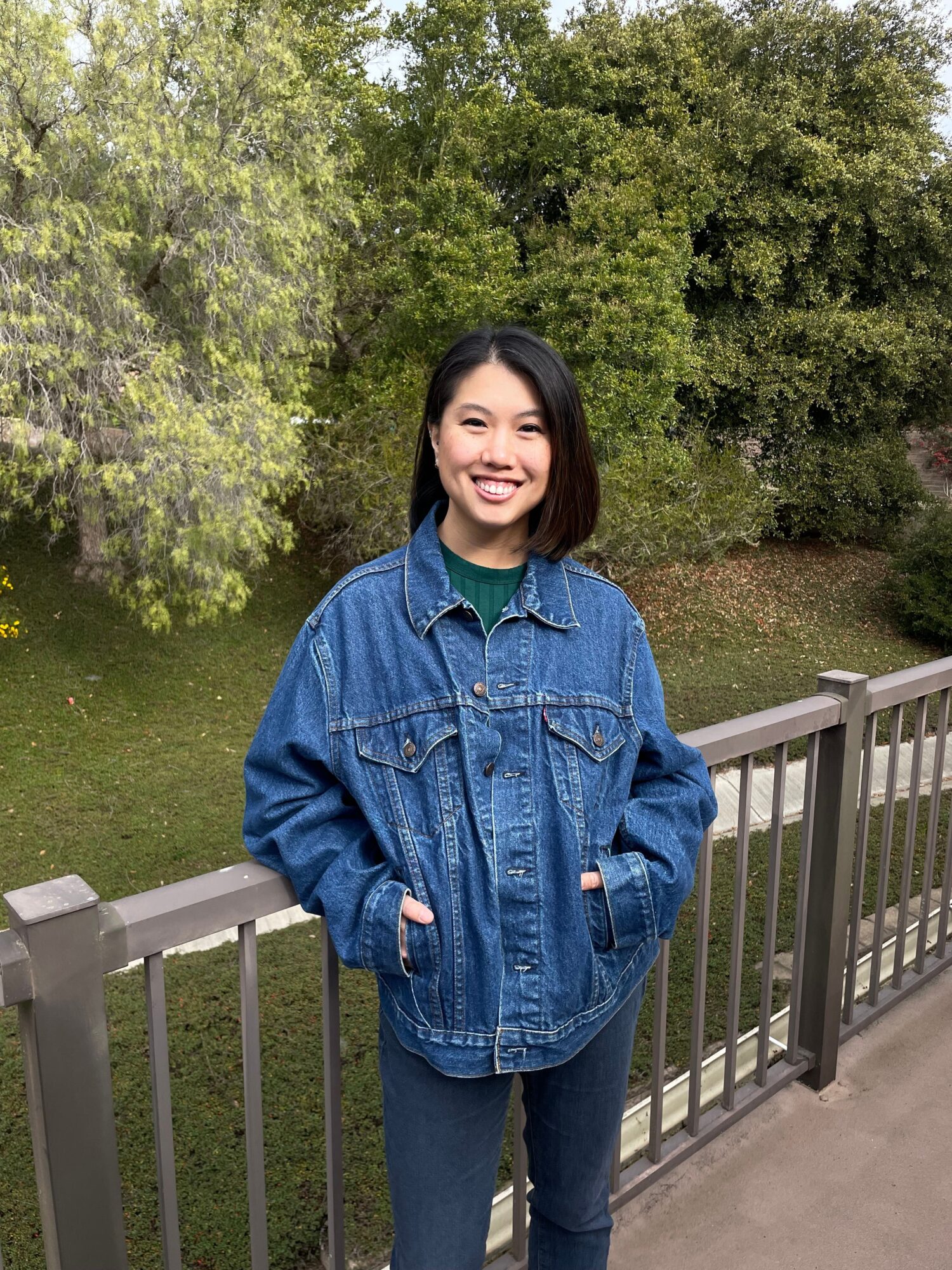 Woman with dark hair smiling, wearing a denim jacket and jeans, standing outdoors near a railing with trees in background.