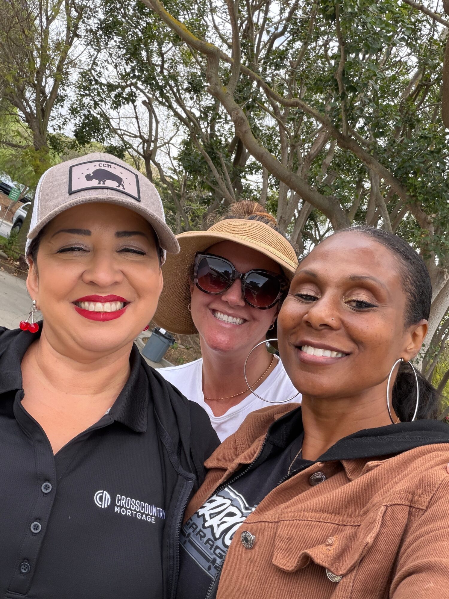 Three women smiling outdoors with trees in the background, one wearing a hat and sunglasses, all dressed casually.