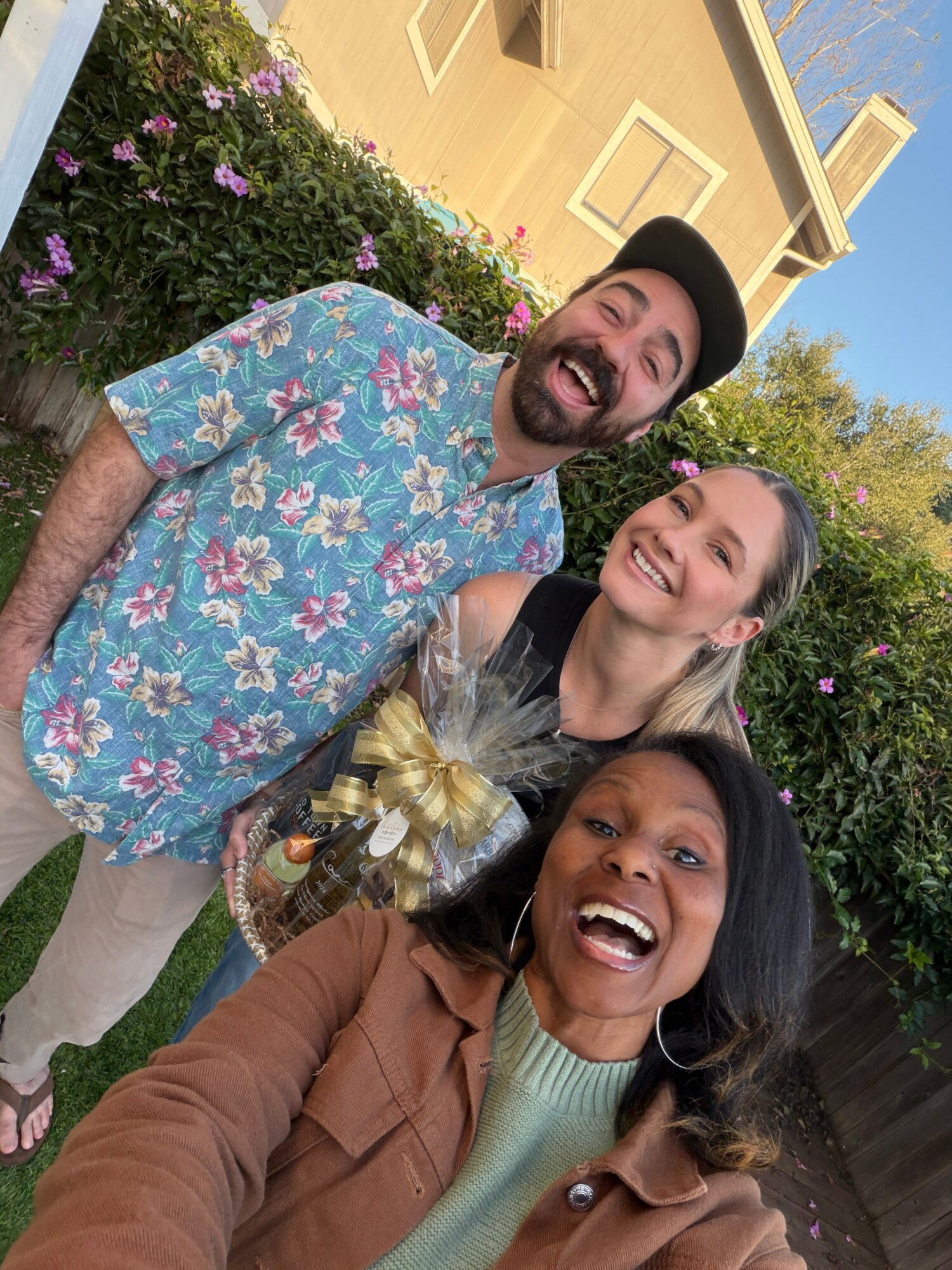 Three people smiling outdoors with flowers and a house in the background.