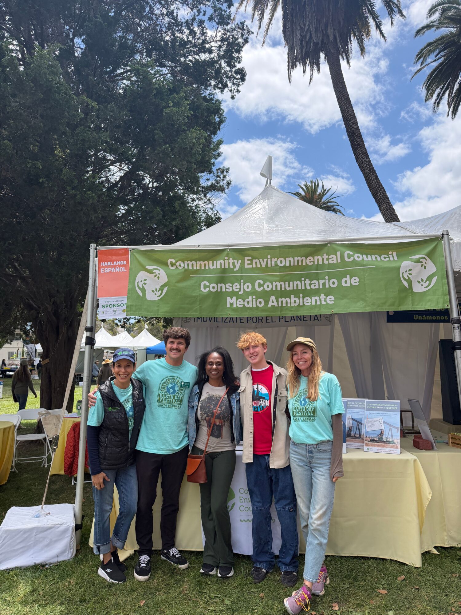 Group of four people standing in front of a green environmental banner at an outdoor event, with trees and a blue sky in the background.