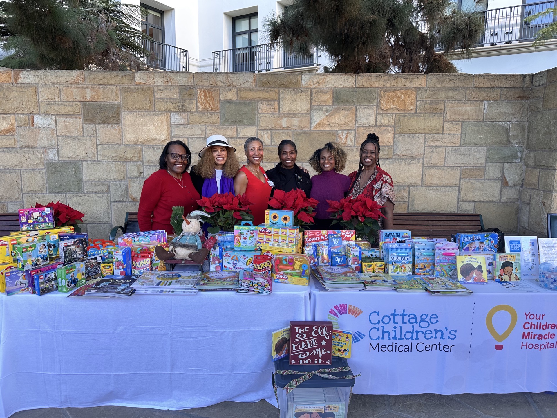 Five women stand behind a table with toys and gifts, outdoors against a stone wall, with a banner for Cottage Children's Medical Center.