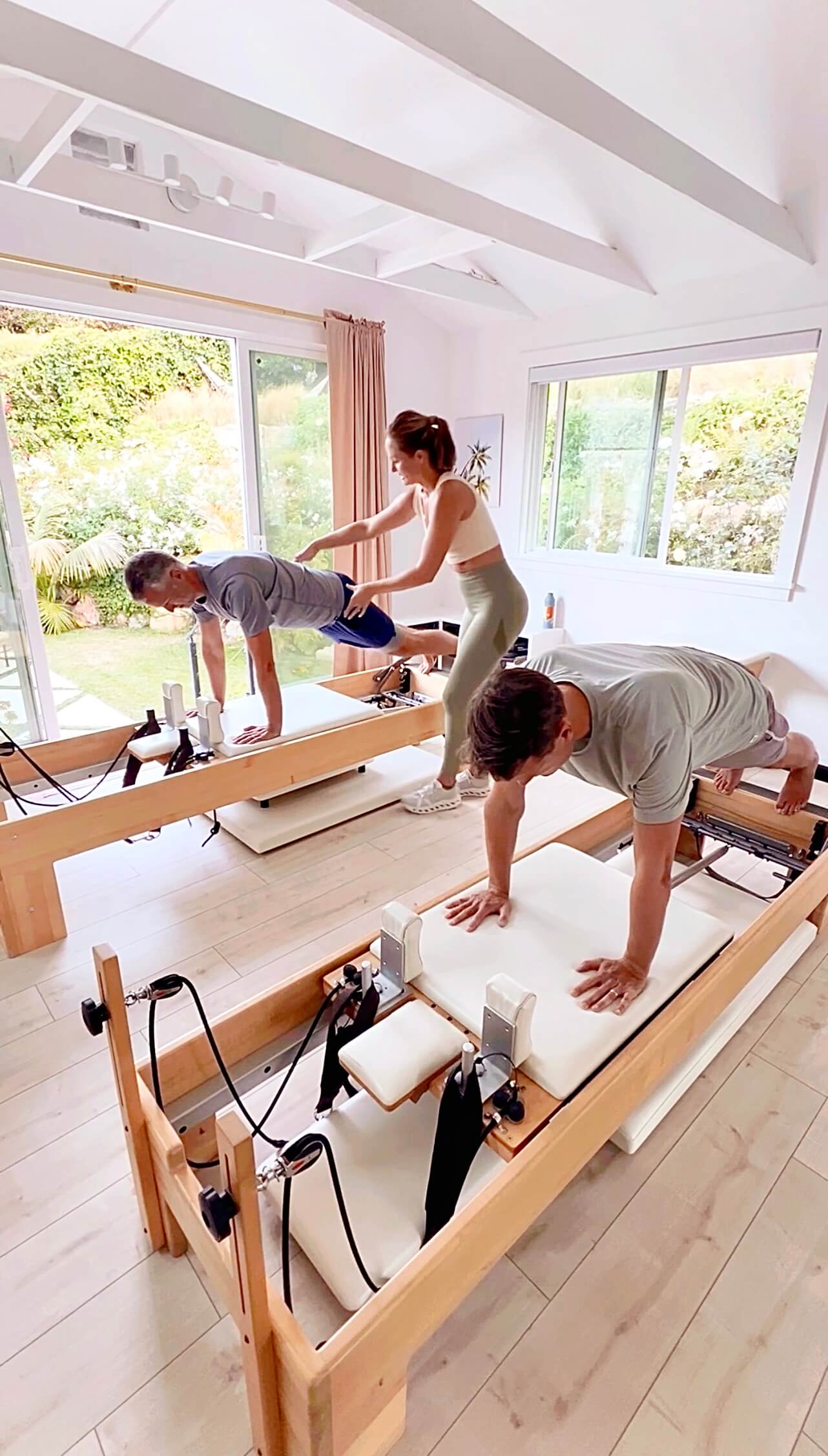 Two people are exercising on Pilates reformers in a bright room with large windows and greenery outside.