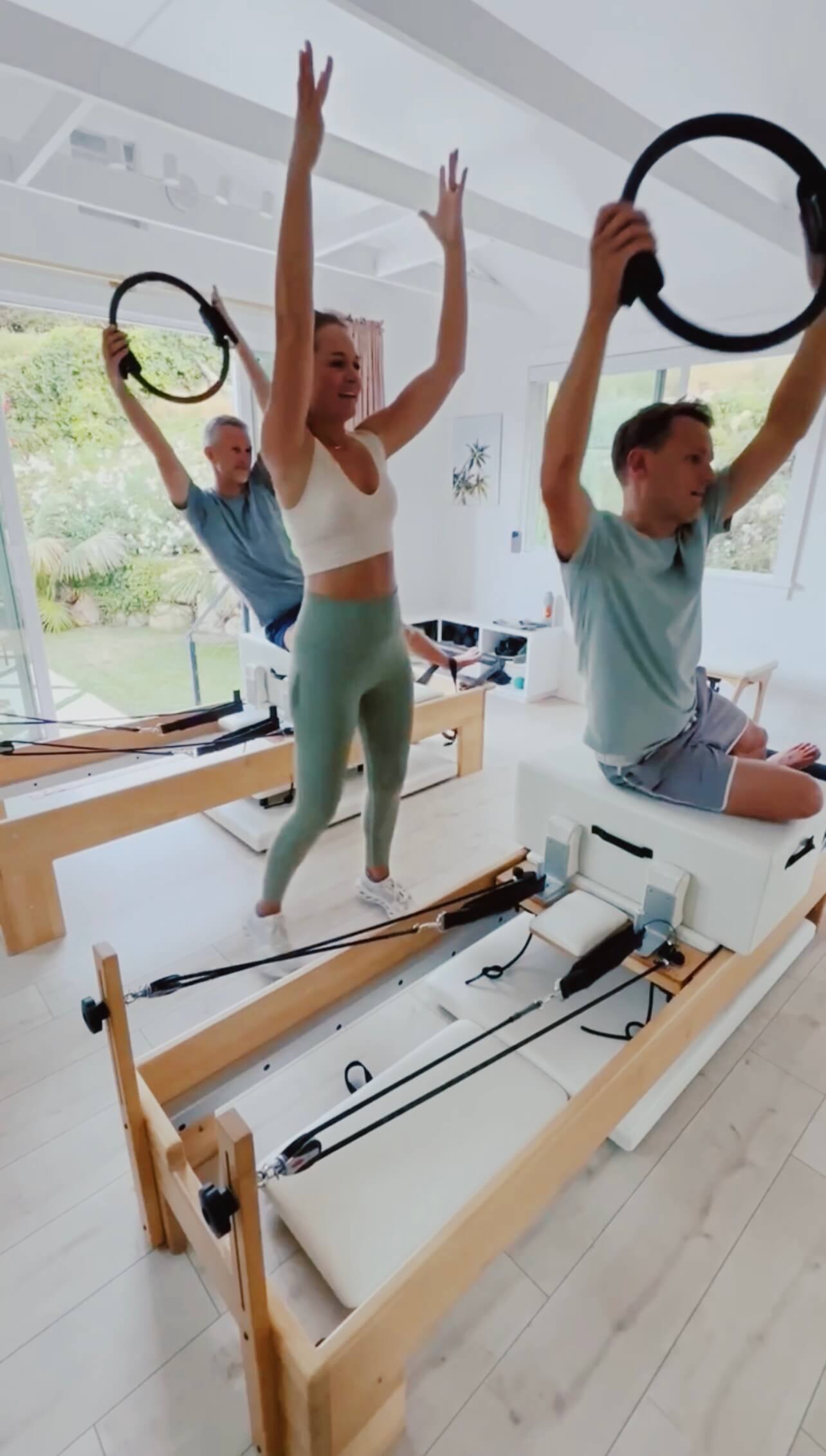 Three people in a Pilates studio holding rings above their heads, with a reformer machine in the foreground.
