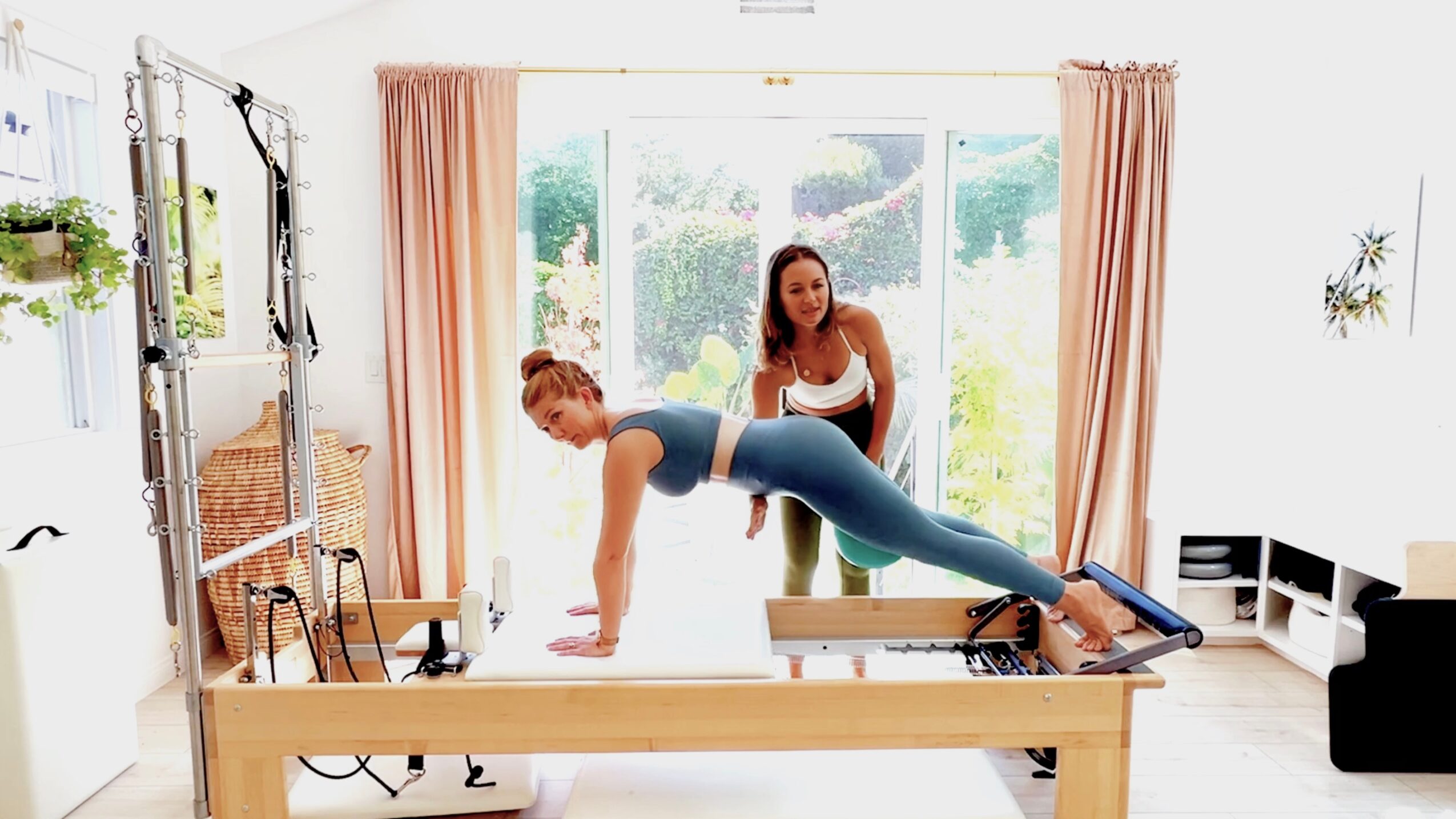 Two women exercising in a bright room with large windows, one on a Pilates reformer and the other assisting.