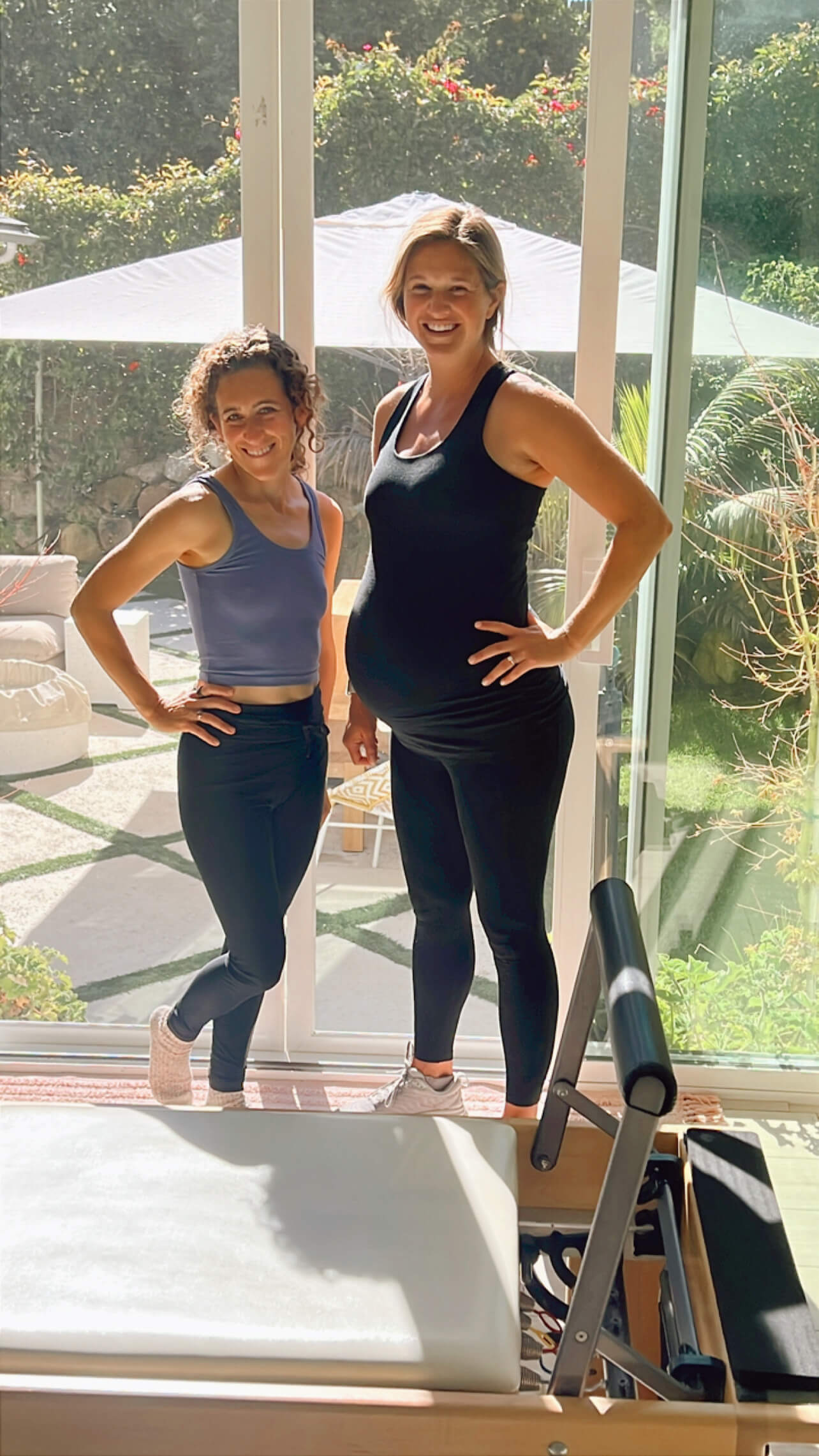 Two women standing indoors near a glass door, smiling, with a garden and umbrella outside in the background.