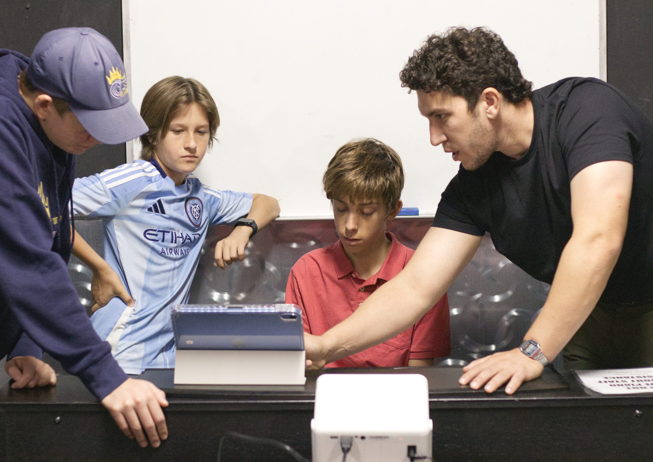 A man and three children are gathered around a table, looking at a device or screen, indoors.