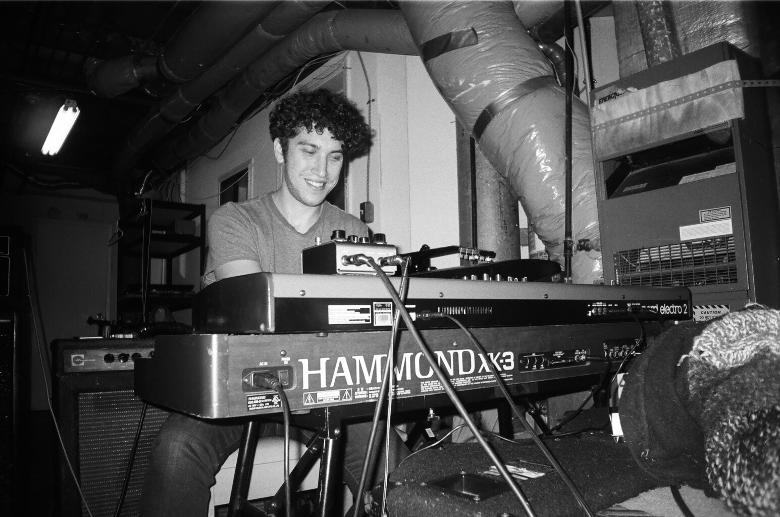 Young man playing keyboard in a room with industrial pipes, smiling, with equipment around him.