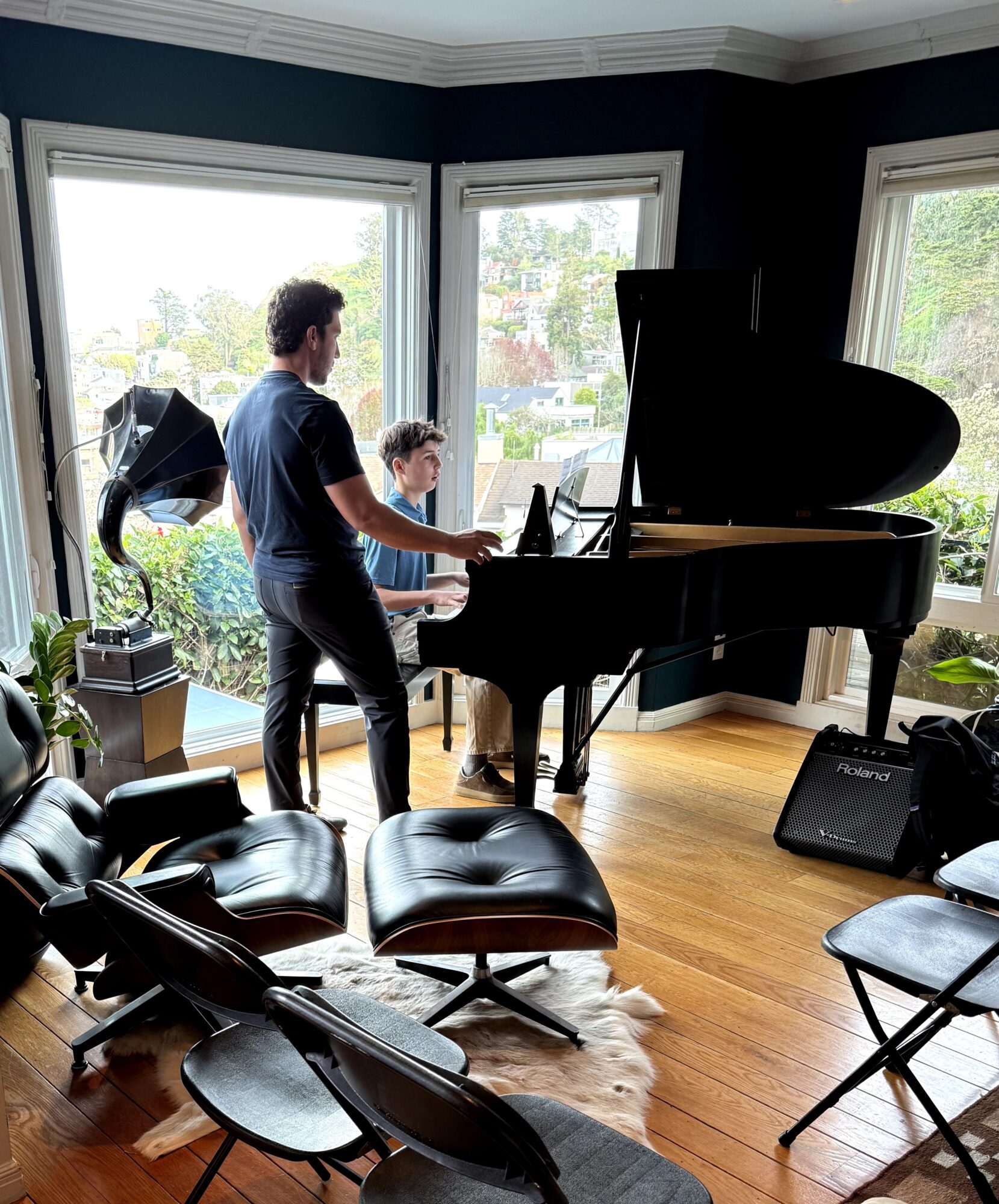Two people, a man and a child, playing piano in a room with large windows and wooden floor, sunlight outside.
