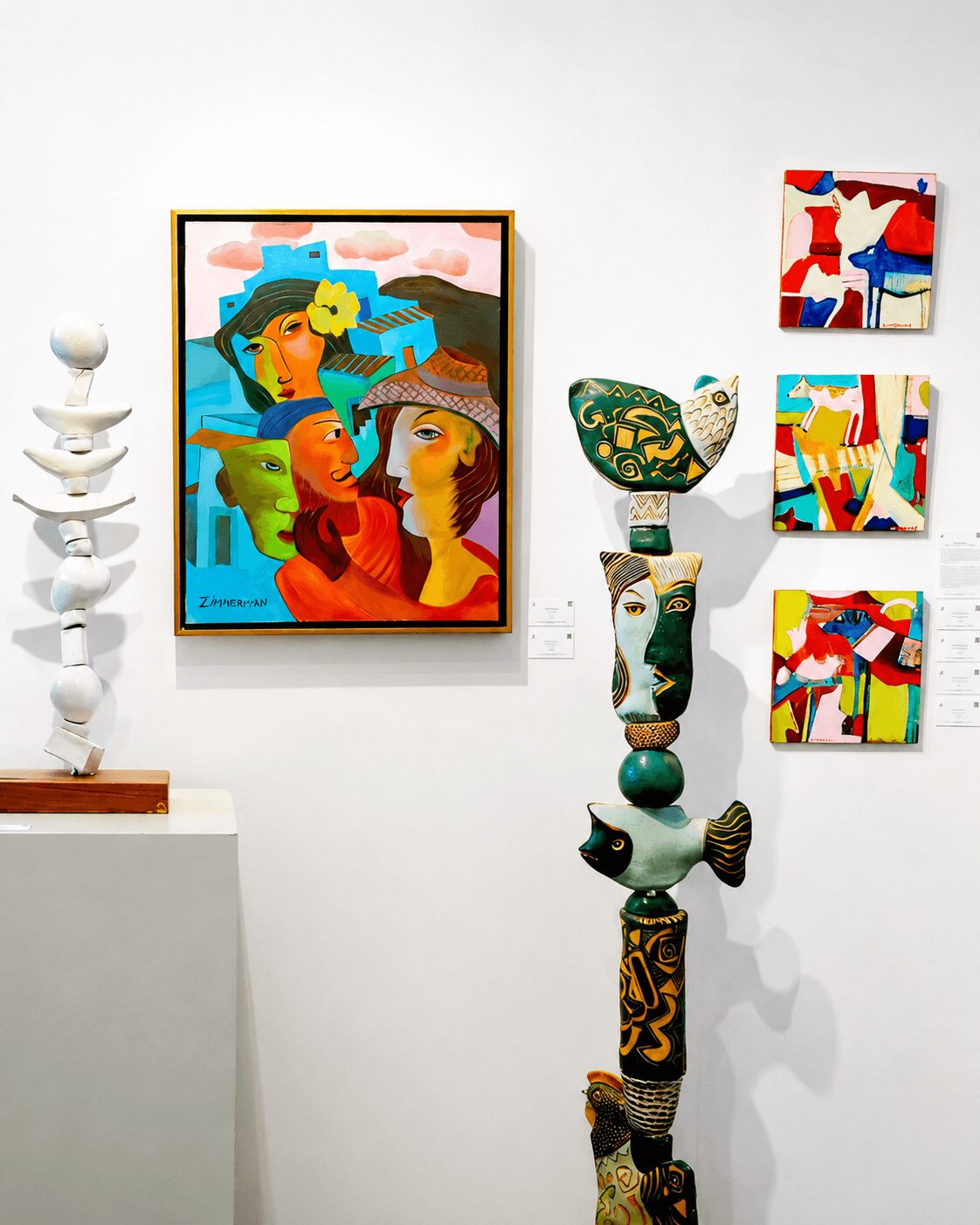 Gallery wall with colorful abstract paintings and sculptures, including a large framed artwork and smaller pieces, on a white wall.