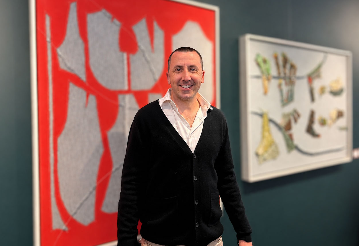Man smiling in art gallery with abstract paintings on the wall behind him.