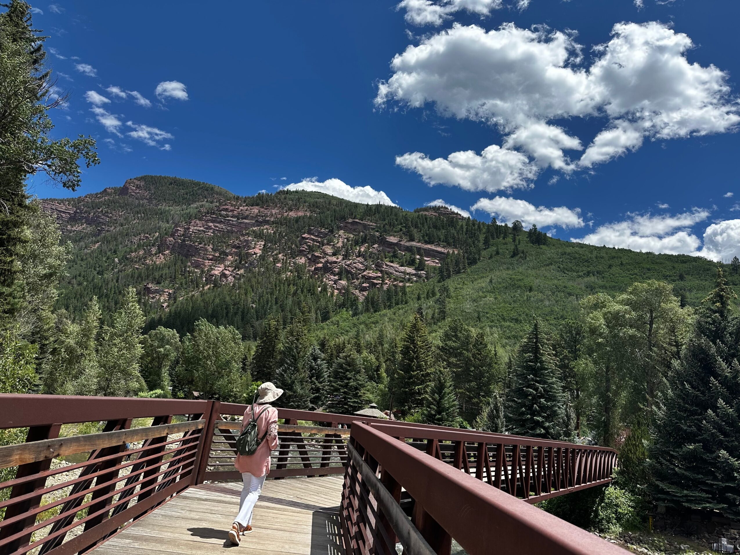 Person walking on a wooden bridge with green mountains and blue sky in the background.