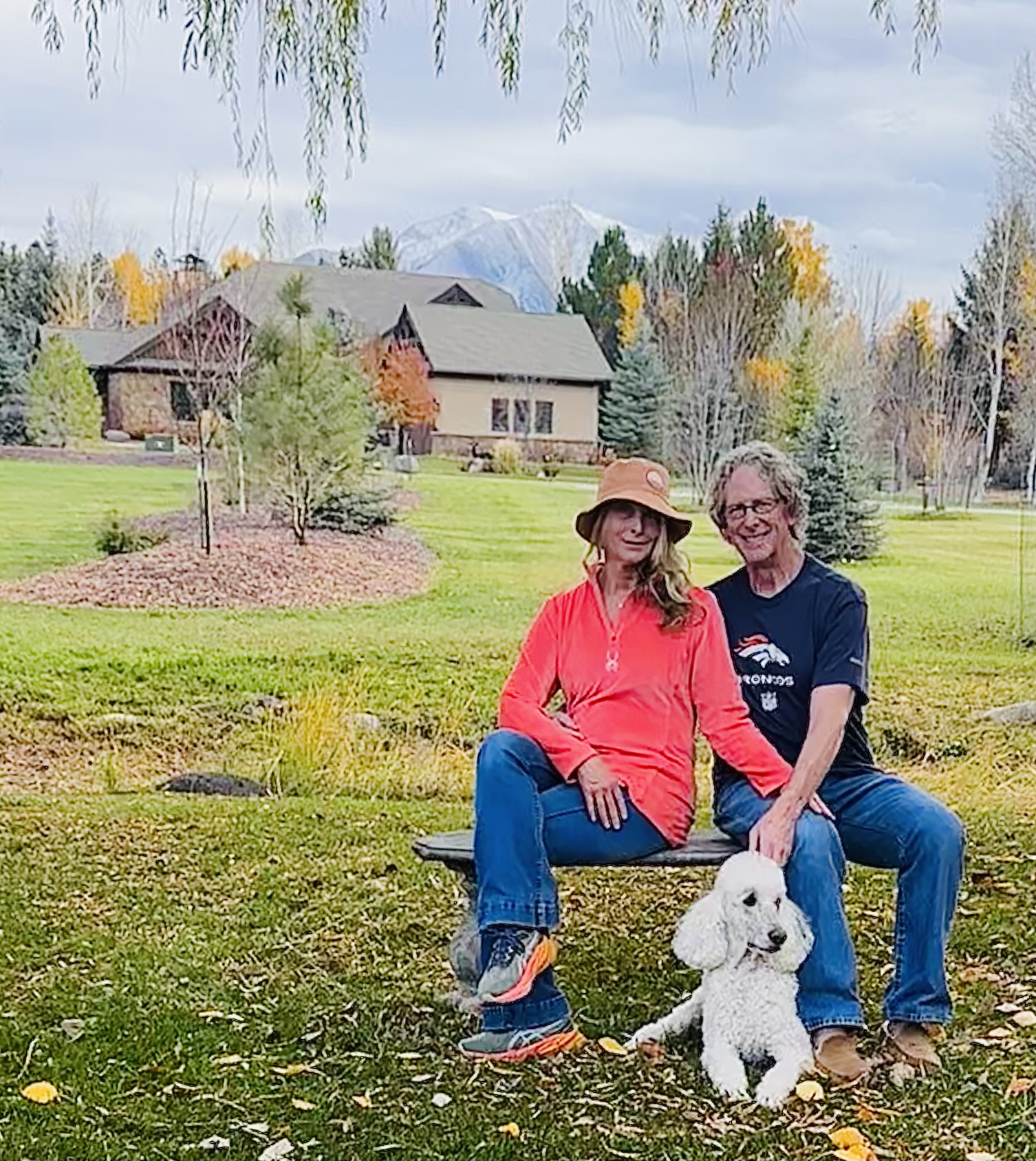 Two people sit on a bench outdoors with a white dog, trees, and houses in the background, under a cloudy sky.