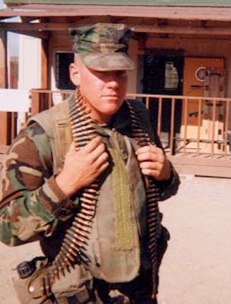 Young person in military uniform holding a bandolier, standing outdoors in front of a wooden building.