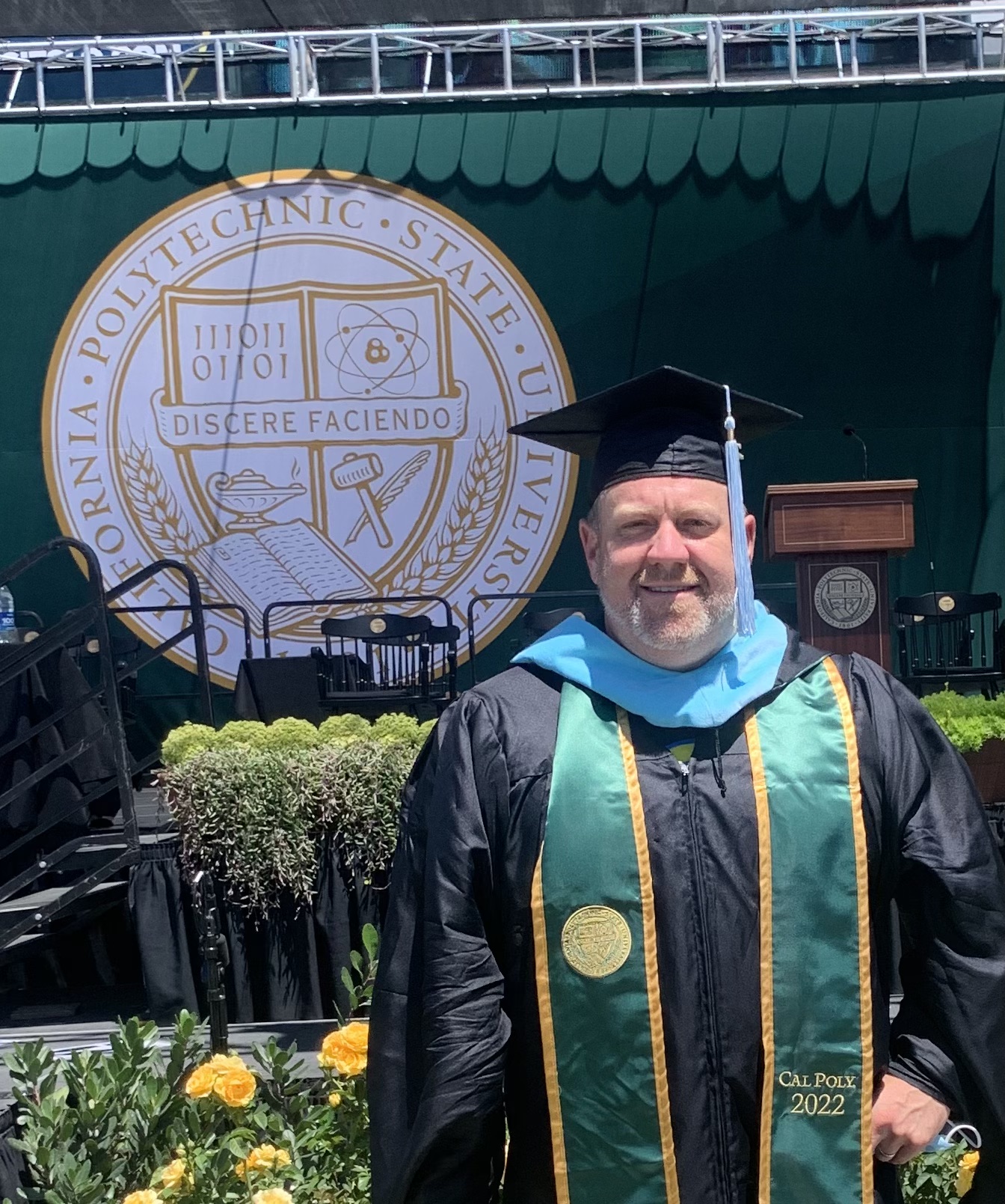 Graduate in cap and gown standing outdoors with university emblem in background, smiling, during graduation ceremony.