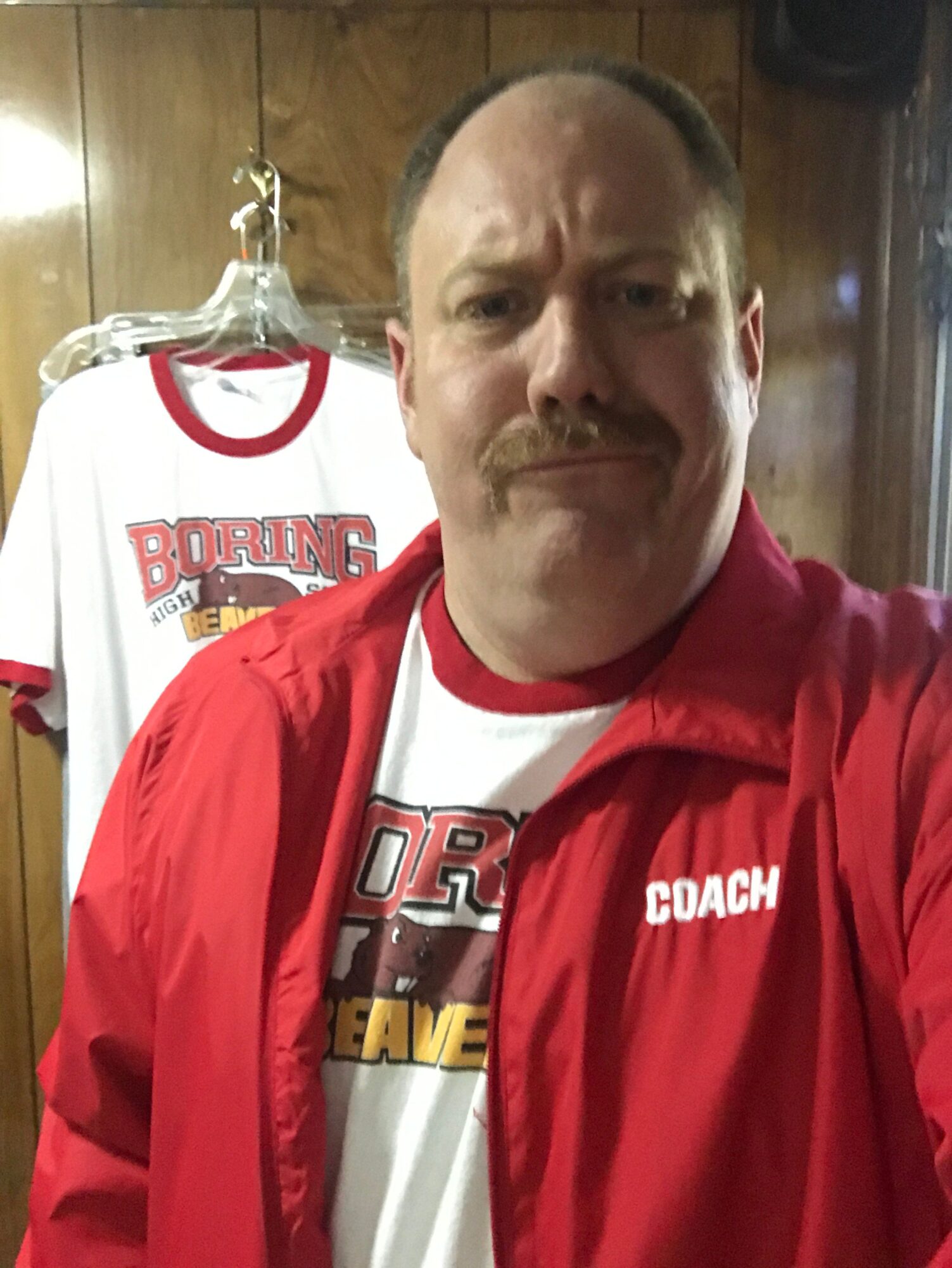 Man with a mustache smiling, wearing a red jacket with 'COACH' on it, standing indoors with a wooden background.