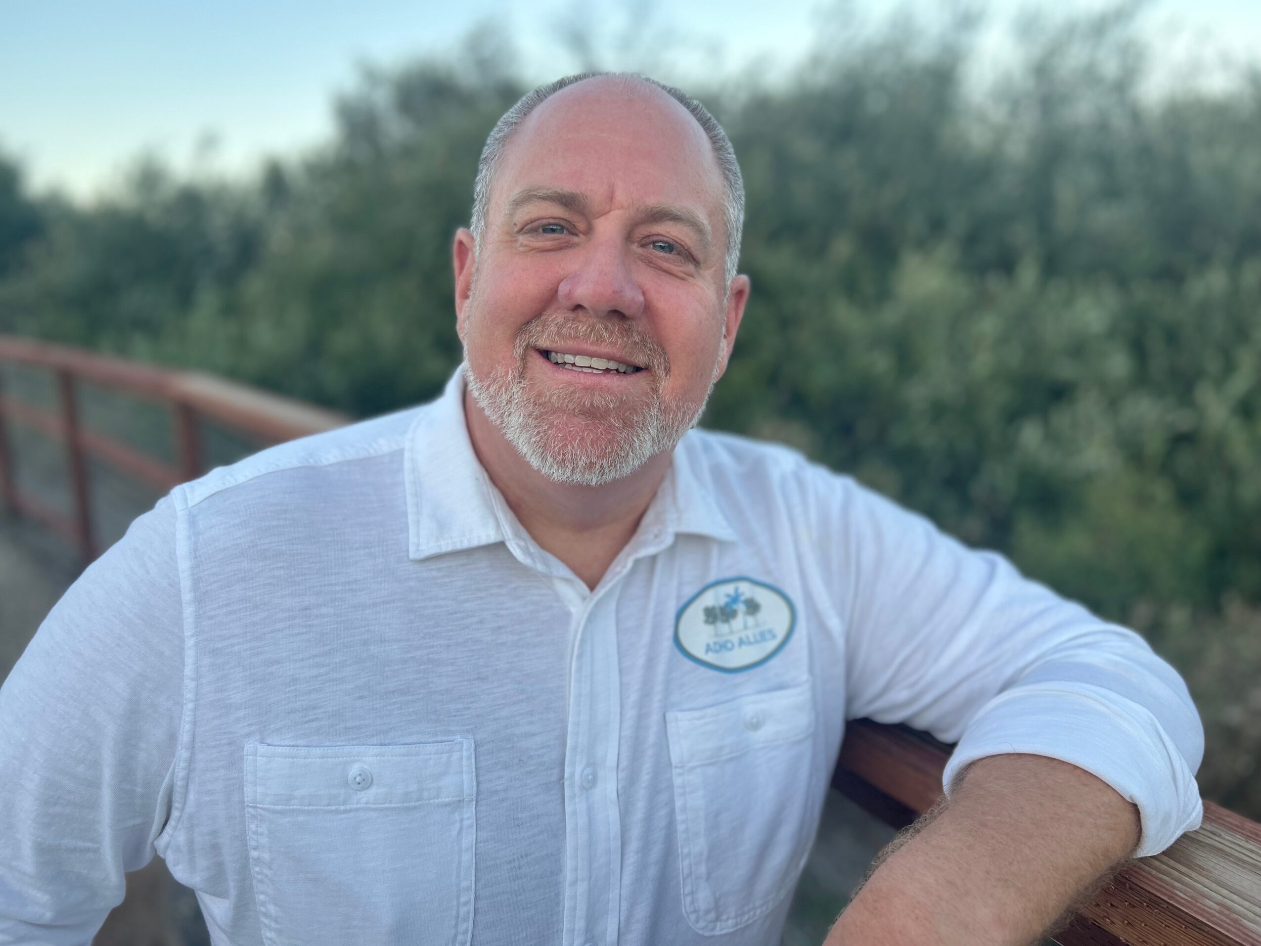 Man with a beard smiling outdoors, wearing a white shirt with a name tag, leaning on a wooden railing.