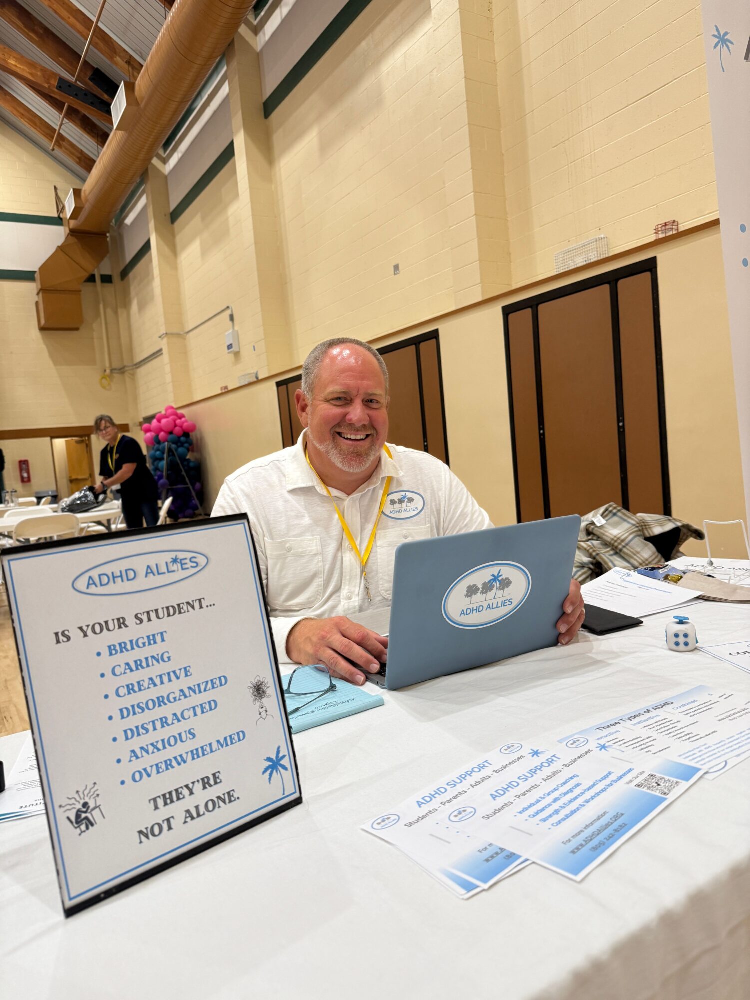 A man in a white shirt sits at a table with a blue box and a sign, smiling at the camera in an indoor setting.