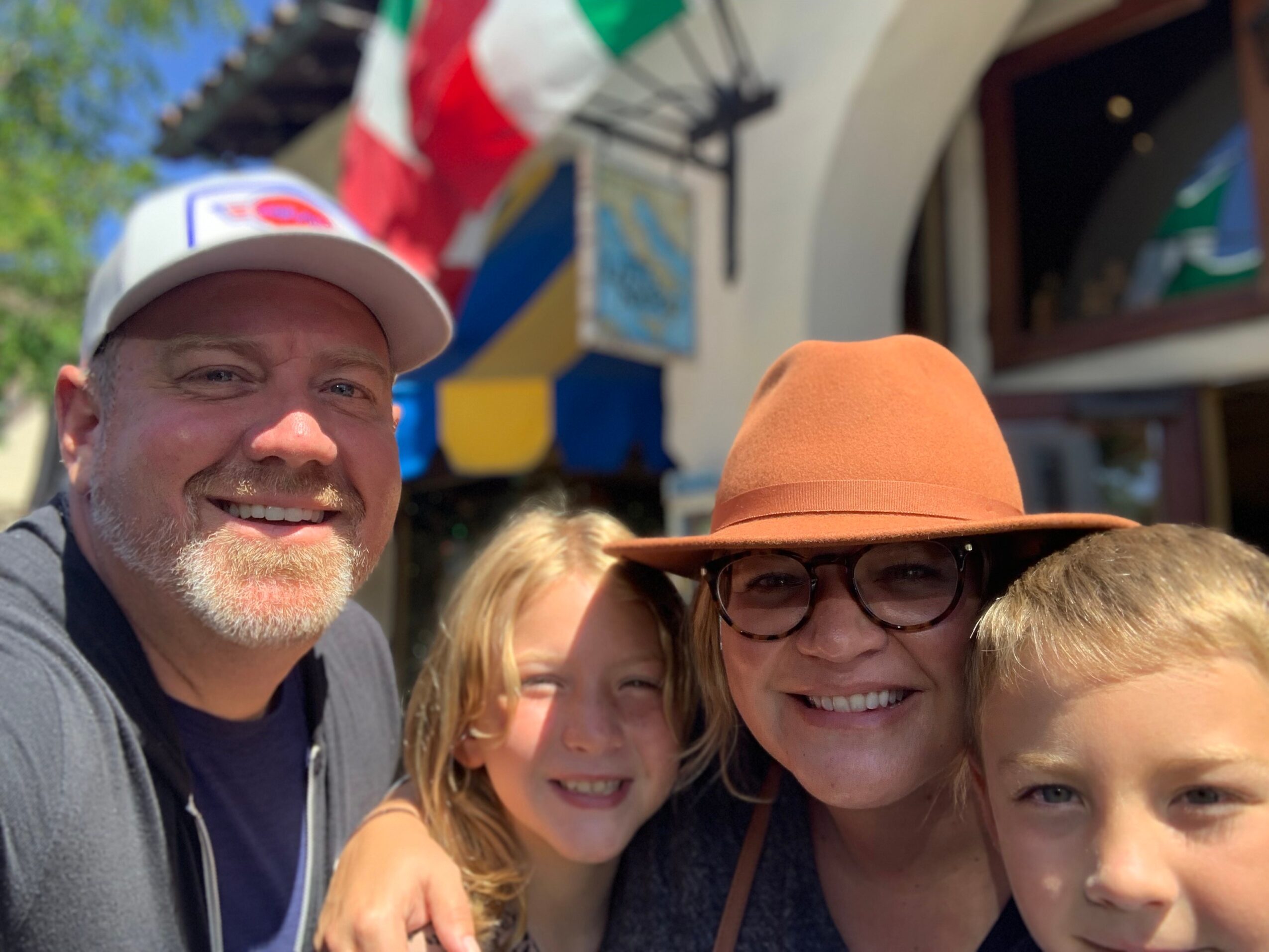 Group of four smiling people outdoors, two adults and two children, with colorful flags and building in background.