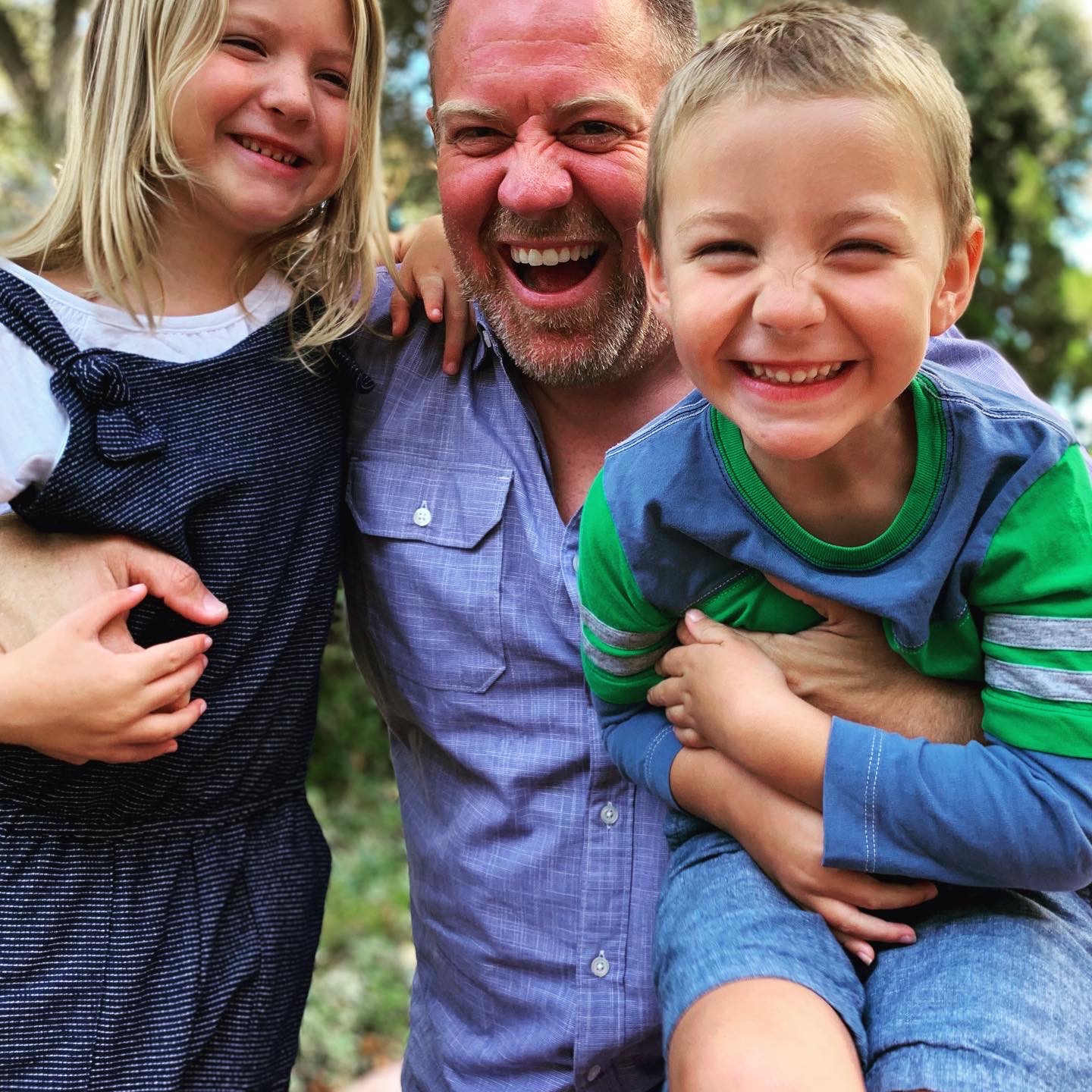 A man with two children outdoors, all smiling and hugging, with trees in the background.