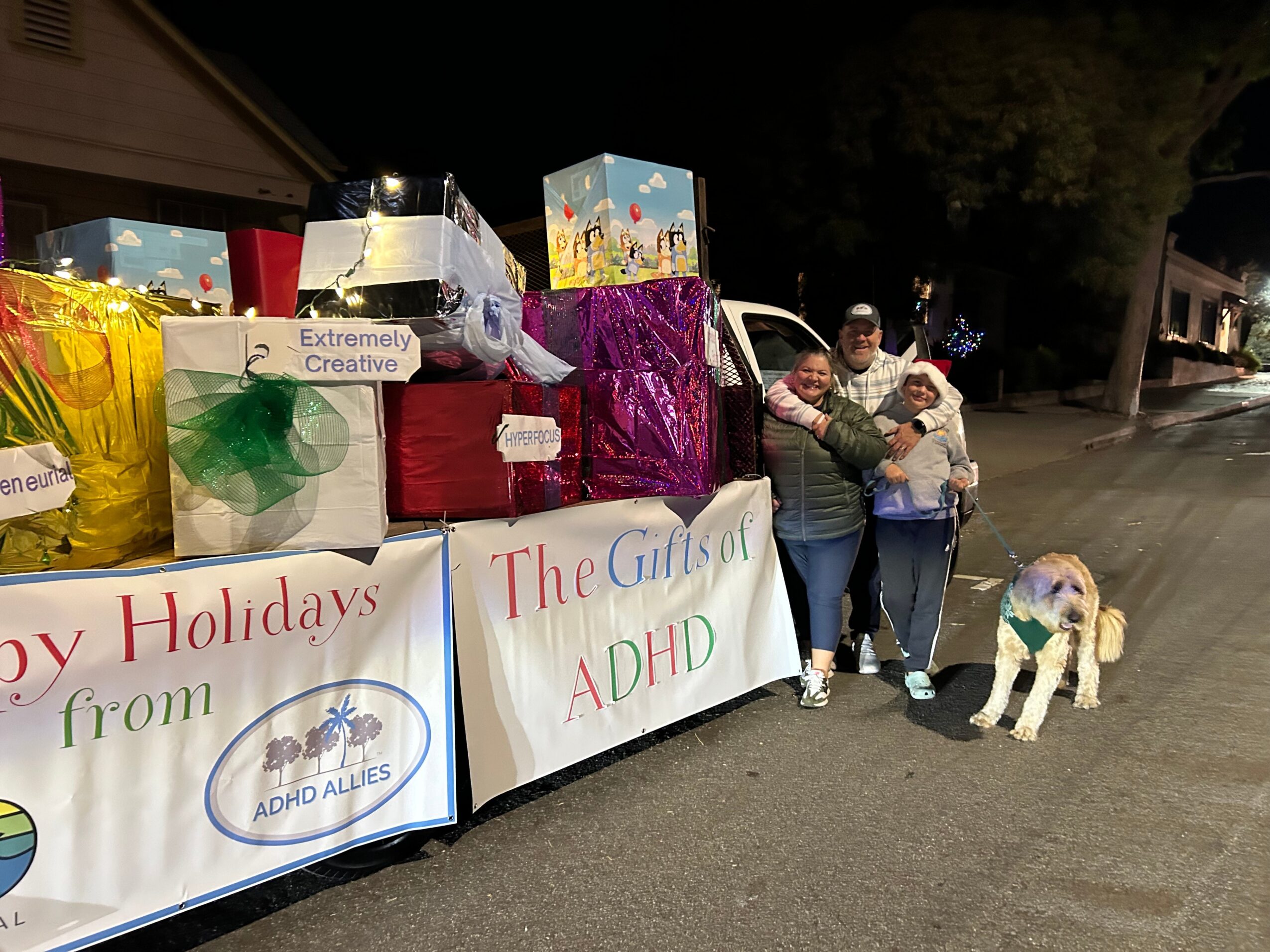 Two people stand next to a table with holiday gift boxes and signs, with a dog on a leash nearby, on a street at night.